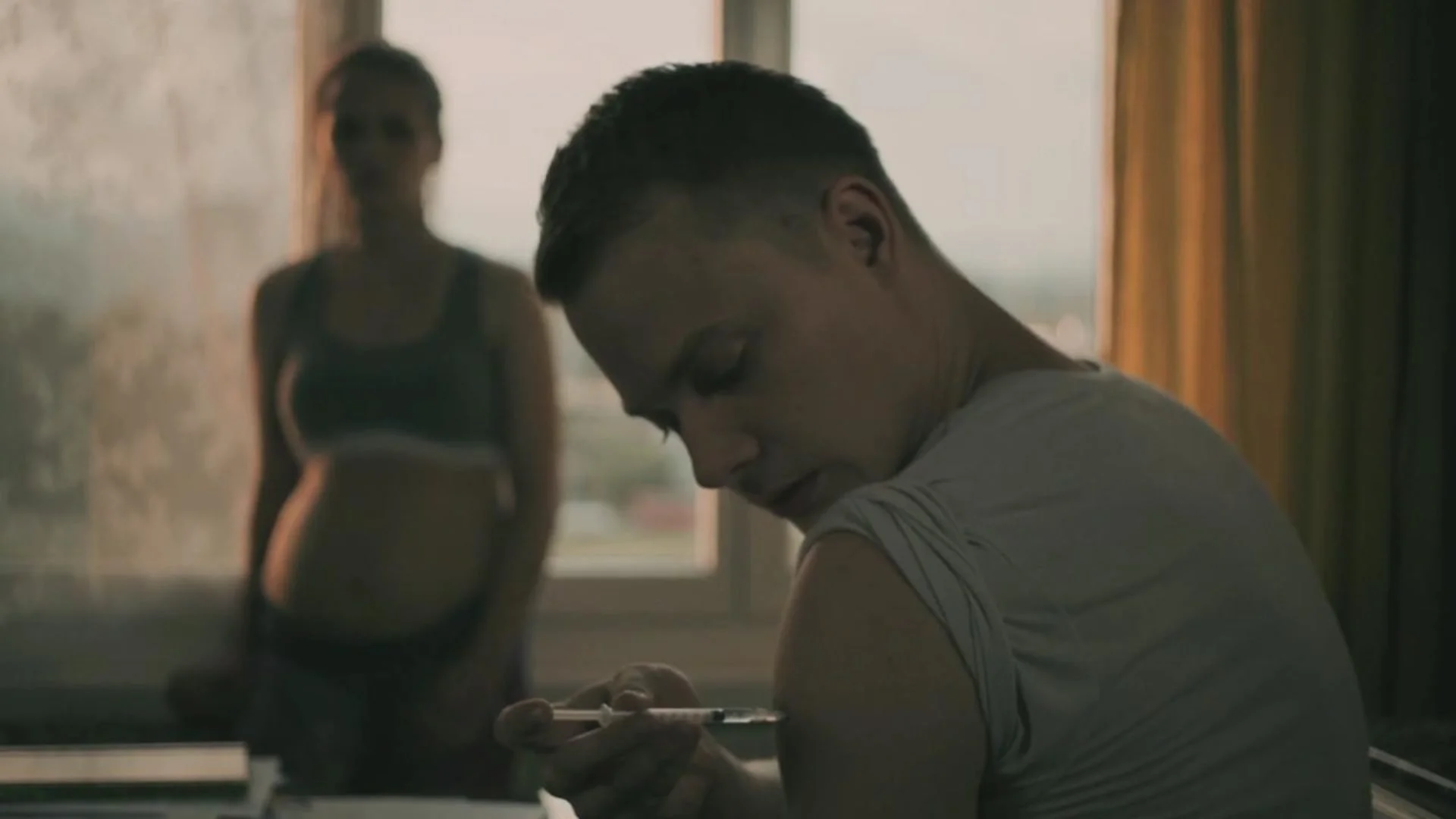 A young man is sitting indoors, looking down at his phone, with a woman in athletic attire standing behind him in front of a window.