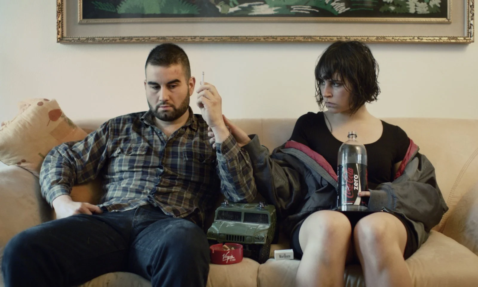 A man and woman sit on a beige couch in a living room. The man is wearing a plaid shirt and appears contemplative. The woman, with short dark hair, wears a black shirt and looks at the man. Multiple items are on the couch, including a toy vehicle, a 