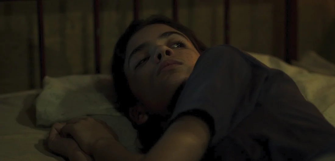 A young boy lying on a bed, resting his head on a pillow, wearing a dark shirt, in a dimly lit room.