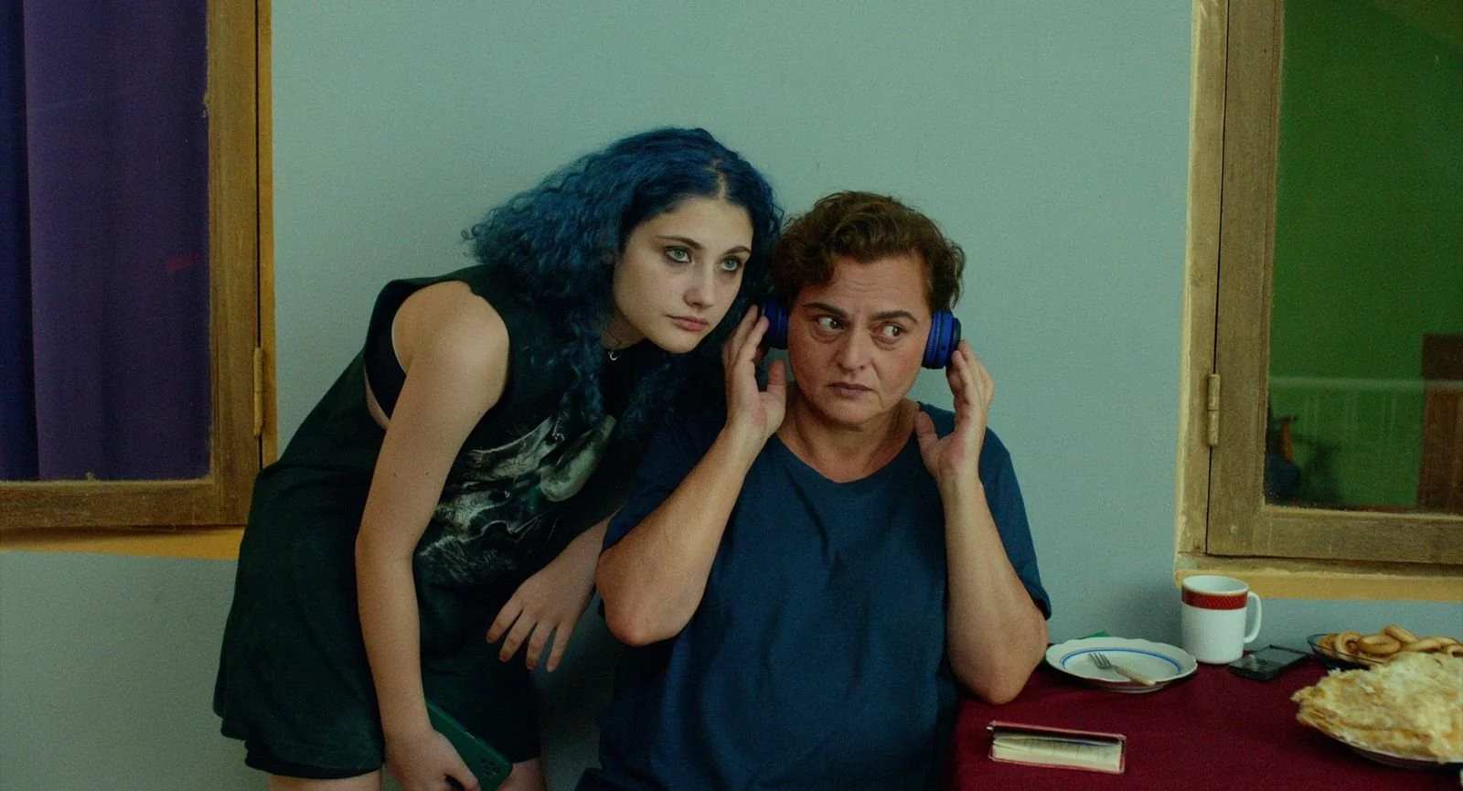 Two women examining music on headphones in a dining room with food and drinks on the table.