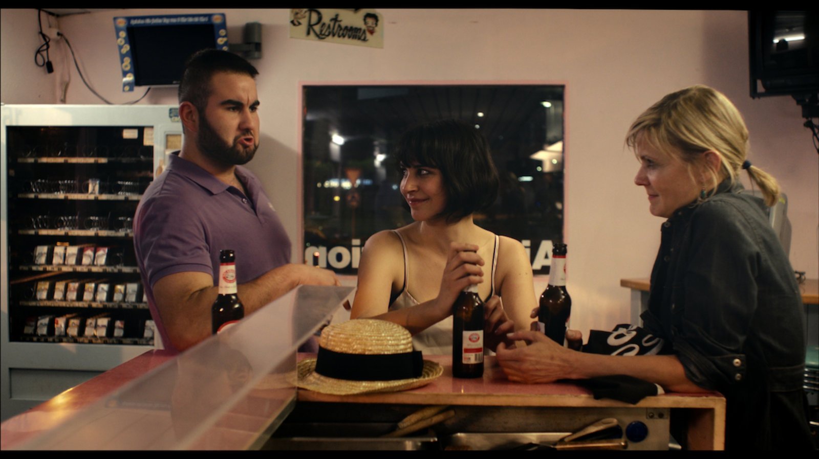 Three people, two women and one man, are talking and holding beer bottles in a casual bar setting. The woman on the left has short dark hair and is smiling, while the woman on the right has blonde hair tied back. A straw hat is on the bar counter, wh