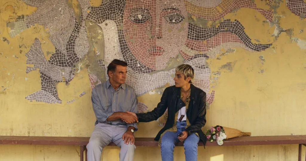 An older man and a younger woman sit on a bench against a worn yellow wall with a mosaic mural of a woman's face. They shake hands, and there is a bouquet of flowers on the bench.