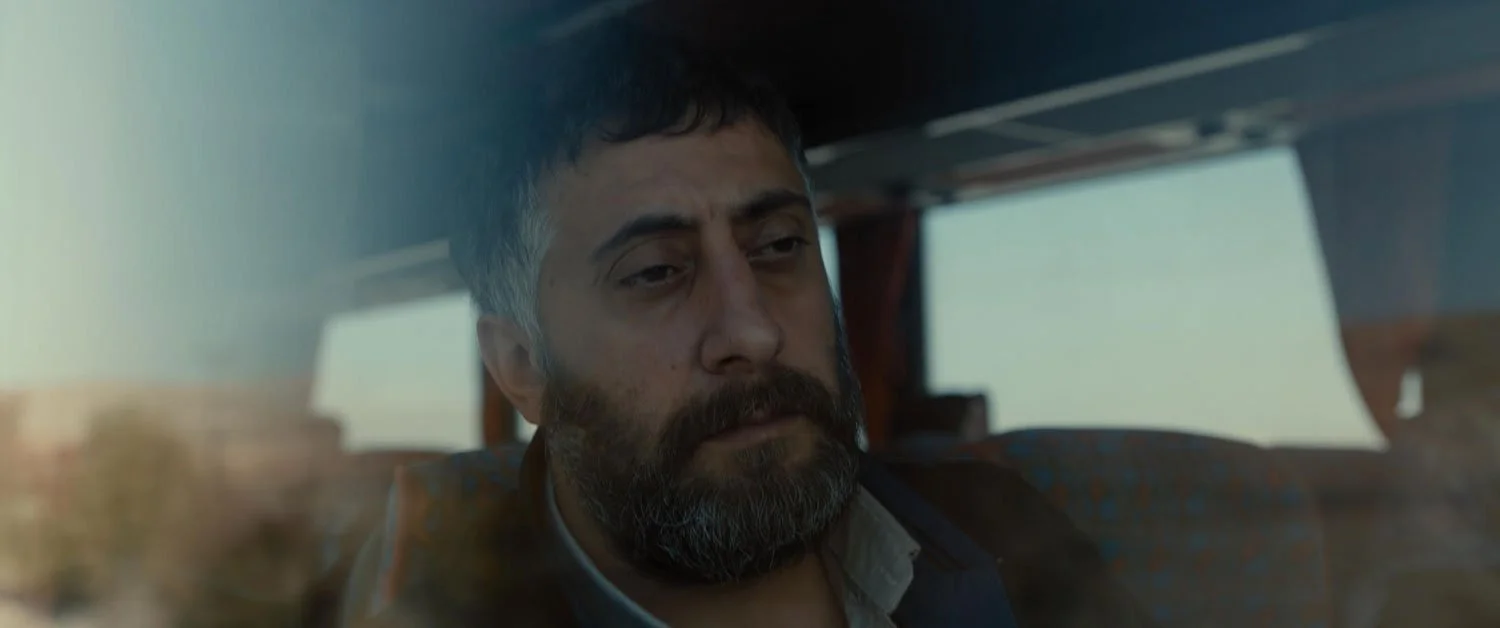 A man with dark hair, a beard, and a mustache, sitting on a bus with a serious expression, looking out the window.
