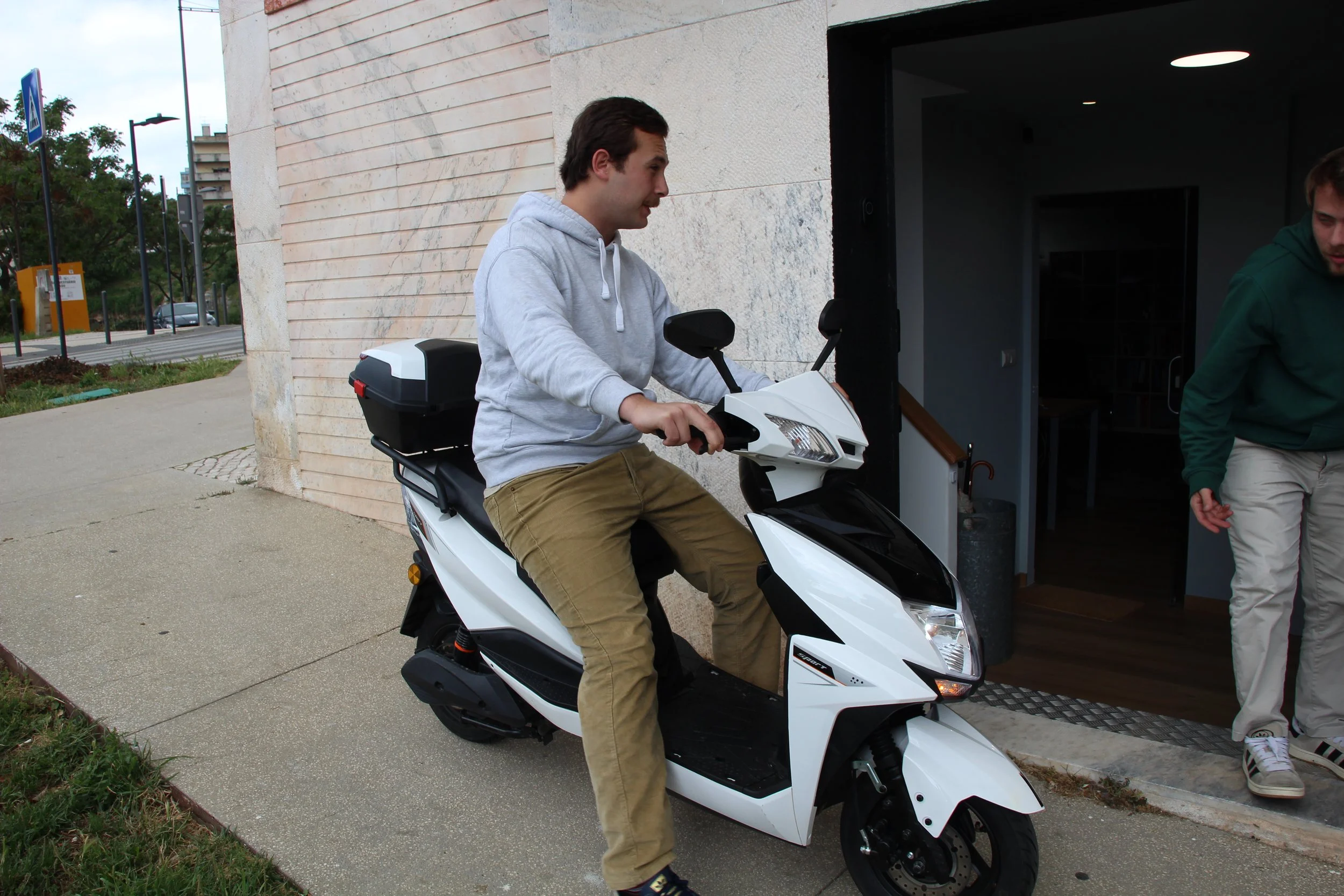 A young man with dark hair wearing a gray hoodie and khaki pants sitting on a white scooter outside a building, with another person partially visible to his right and a background of trees, street signs, and buildings.