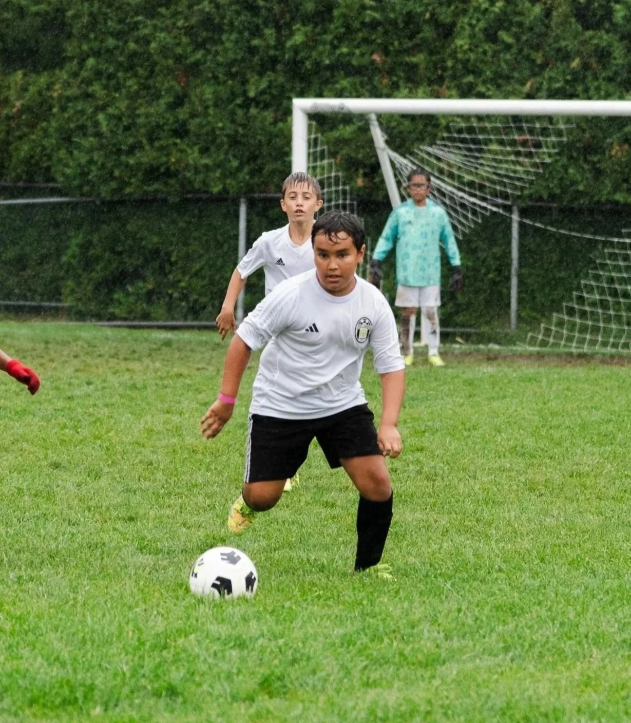 Children playing soccer on a grassy field with a goal in the background, rain falling.