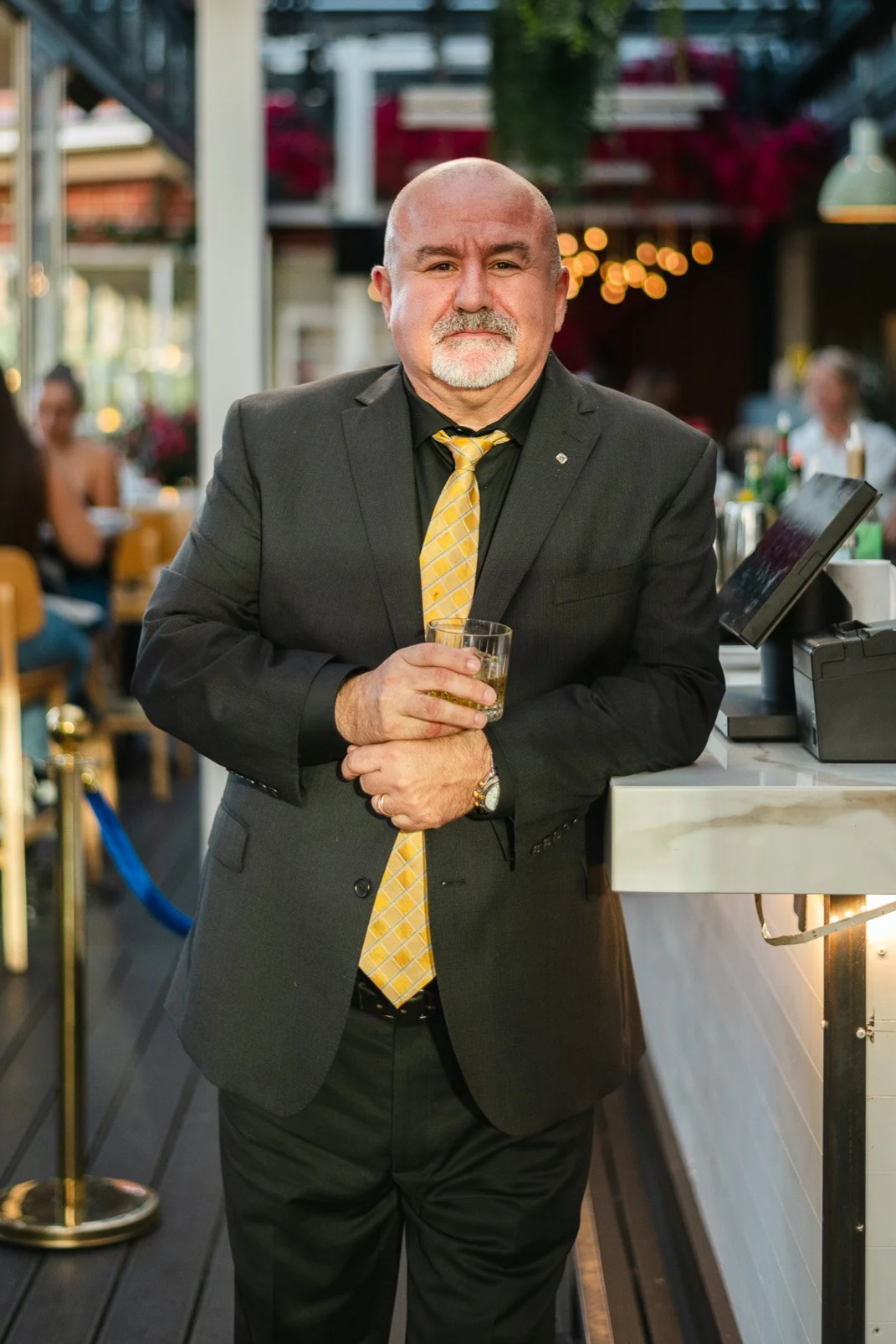 A middle-aged man in a black suit and yellow tie, holding a glass of whiskey, standing at a bar in a restaurant or pub.