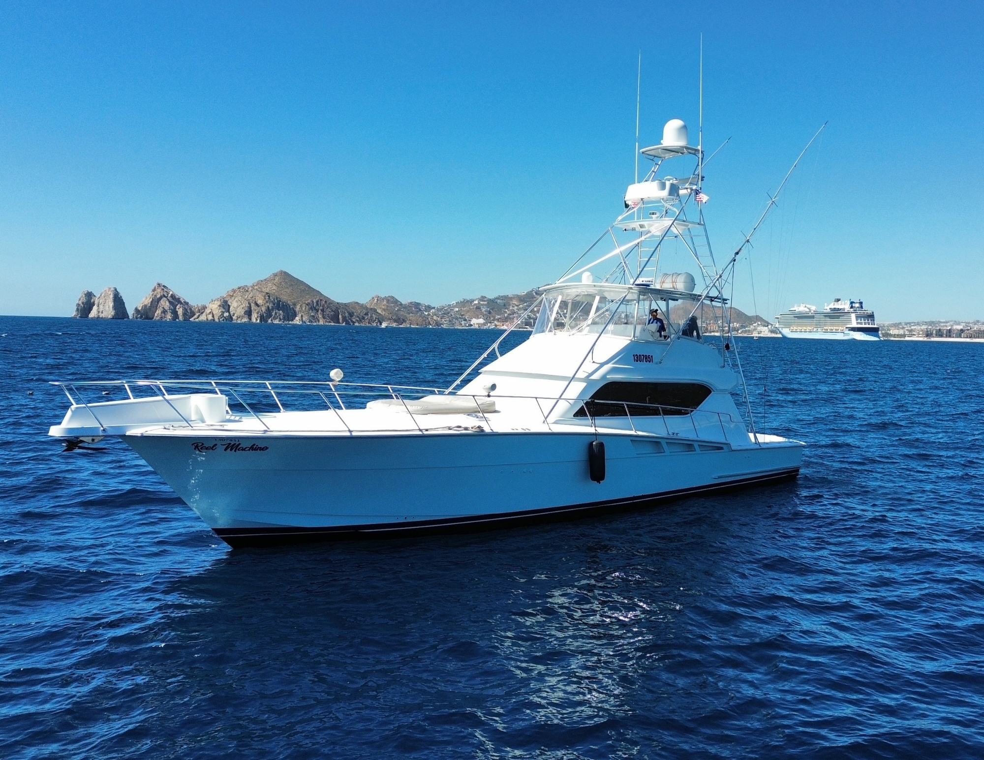 A white yacht named 'Reel Machine' floating on deep blue water with a mountainous coastline in the background under a clear blue sky. Another large cruise ship is visible in the distance.