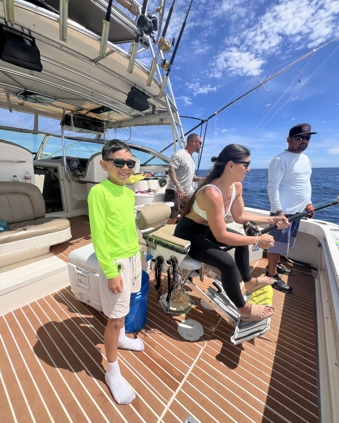 Four people fishing on a boat, with blue sky and ocean in the background. A young boy in a neon green shirt and white shorts is standing, smiling, wearing sunglasses. A woman sitting on a chair is holding a fishing rod. Two men are standing behind her, also fishing, with one wearing a white long-sleeve shirt, sunglasses, and a cap, and the other with tattoos and sunglasses.