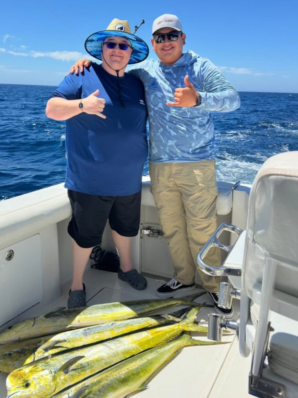 Two men on a boat with yellowfin tuna fish laid out on the deck, smiling and making 'shaka' hand signs, wearing casual and fishing gear, with an ocean backdrop and a clear blue sky.