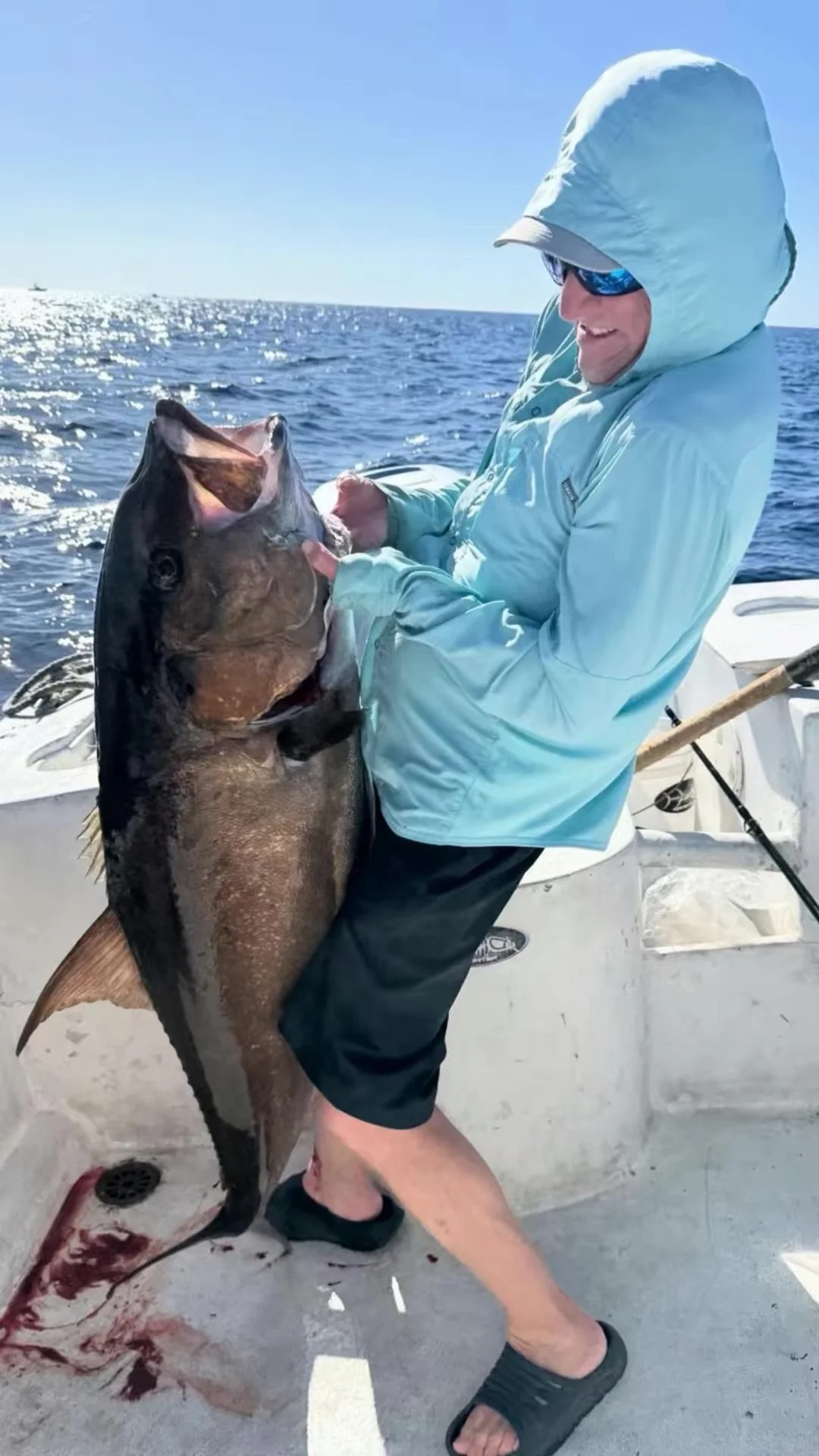 Person in light blue jacket and black shorts holding a large fish on a boat with water and sky in the background.