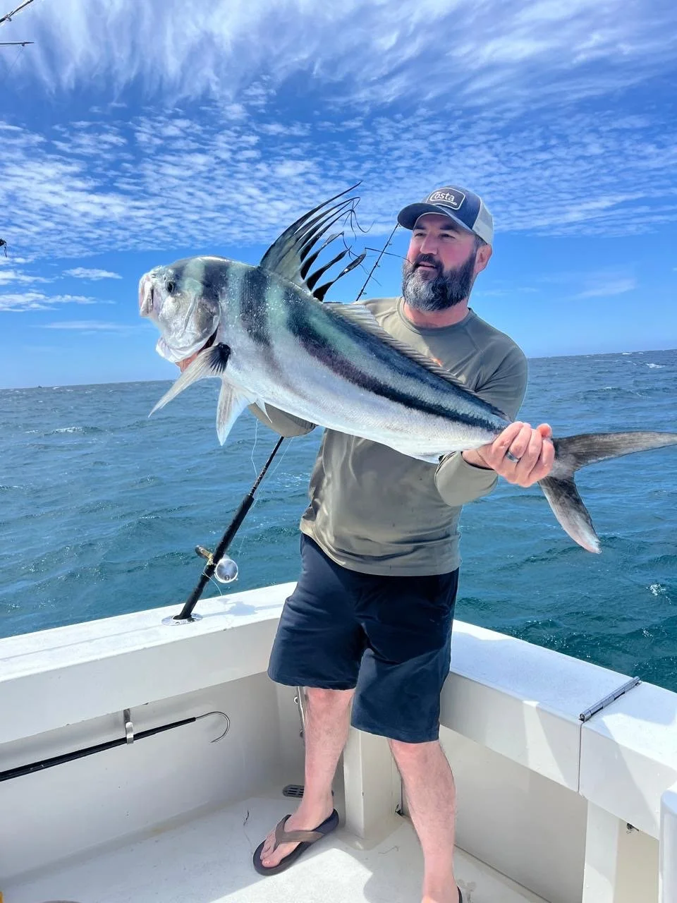 Man holding a large fish on a boat in the ocean under a blue sky with clouds.