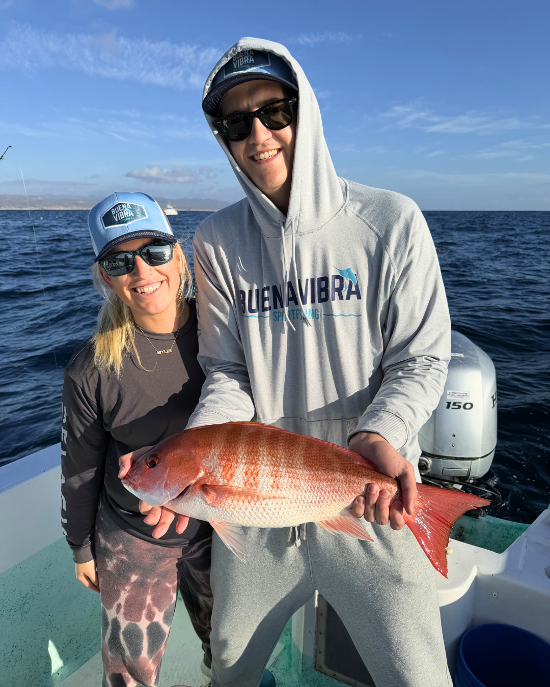 Two people on a boat holding a large red fish, with an ocean and clear sky in the background. The man is wearing a hoodie and sunglasses, and the woman is smiling wearing a cap and sunglasses.