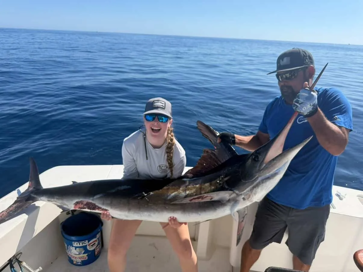 Two people on a boat holding a large fish with an ocean in the background.