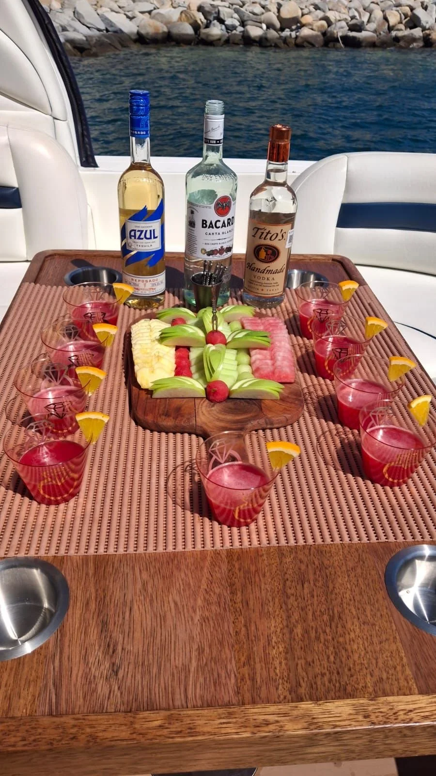 A table set outdoors with a wooden cutting board featuring watermelon, cantaloupe, honeydew, and radishes, surrounded by 12 pink cocktails with lemon wedges on a boat near water with rocks in the background. There are three bottles of alcohol behind the cutting board.