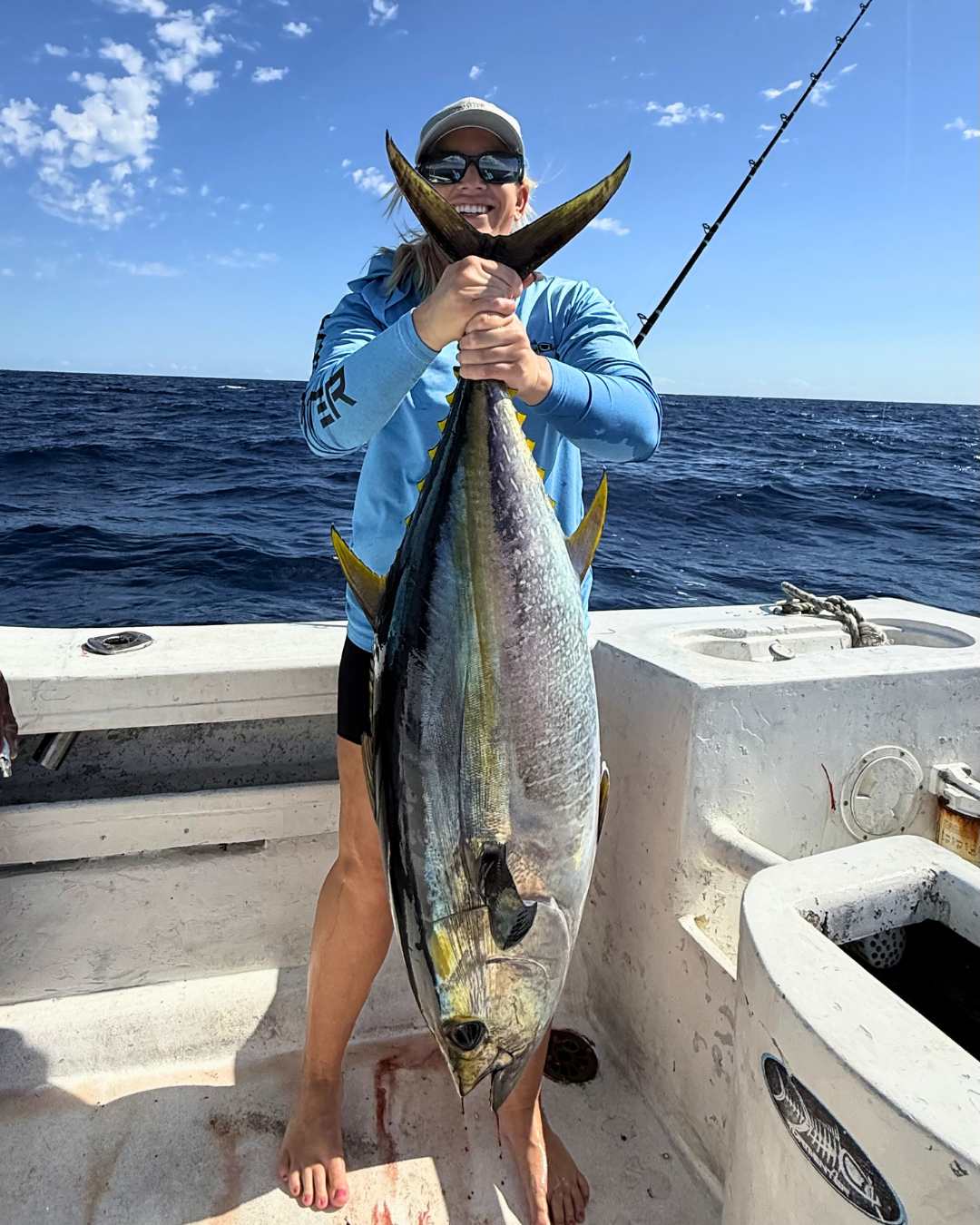 Woman smiling and holding a large yellowtail fish on a boat at sea, wearing sunglasses, a cap, and a blue jacket, with a fishing rod in the background.