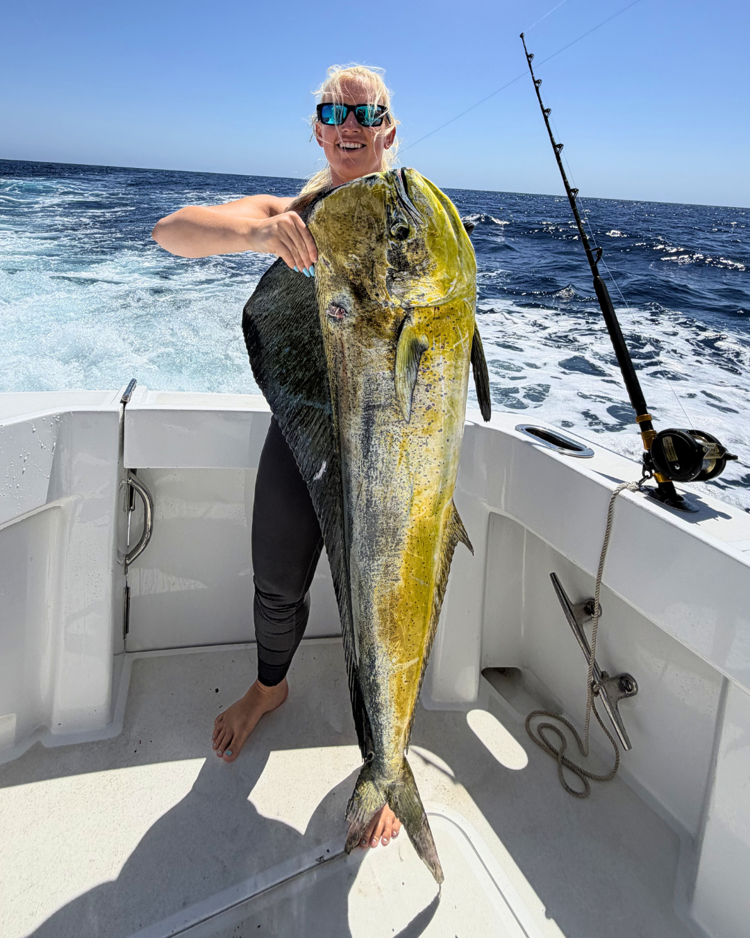 Woman on a boat holding a large fish she caught, with fishing rod and ocean in the background.