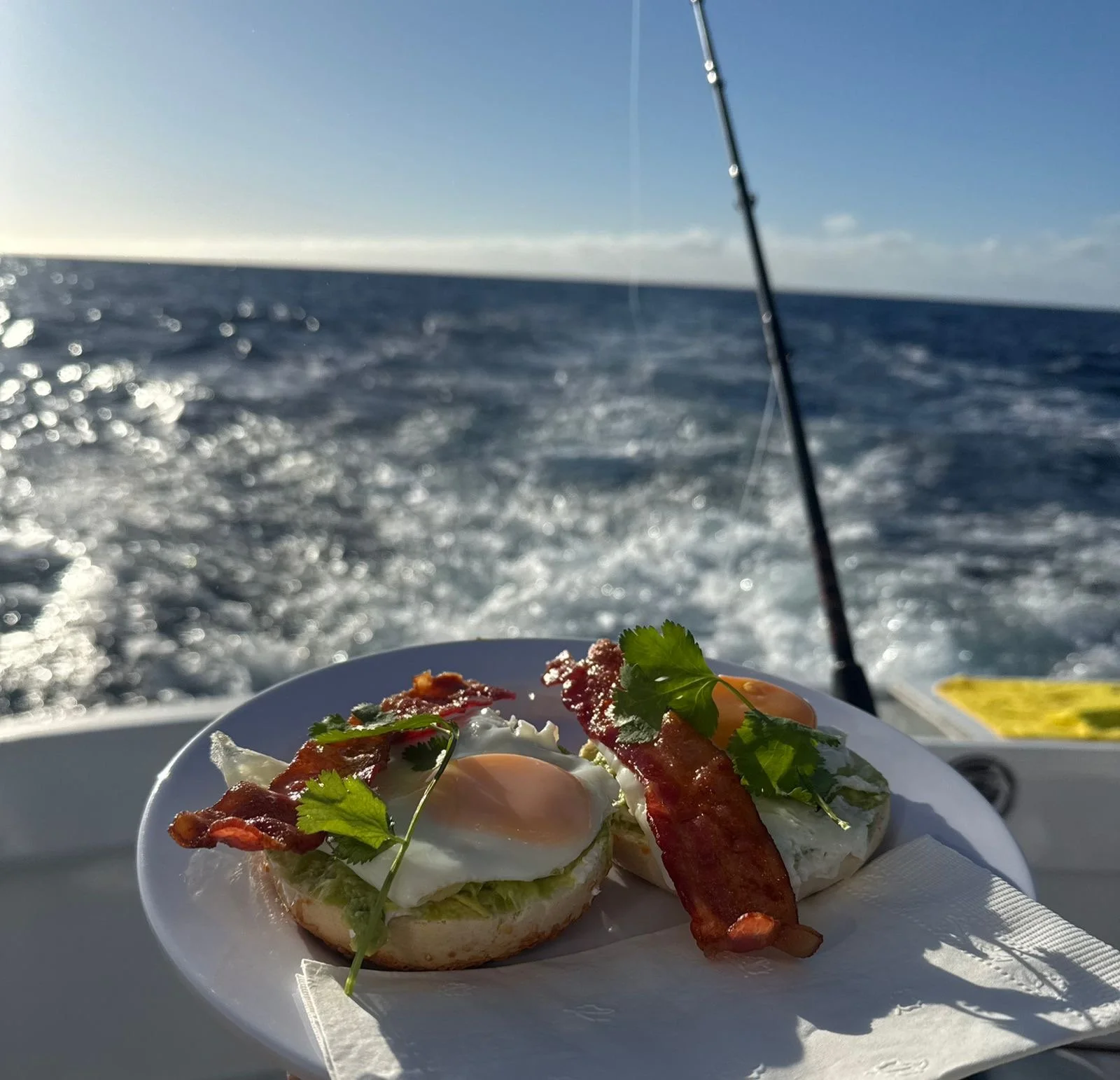 Two servings of eggs Benedict with bacon and cilantro on a white plate, set against the backdrop of the ocean on a boat with a fishing rod visible.