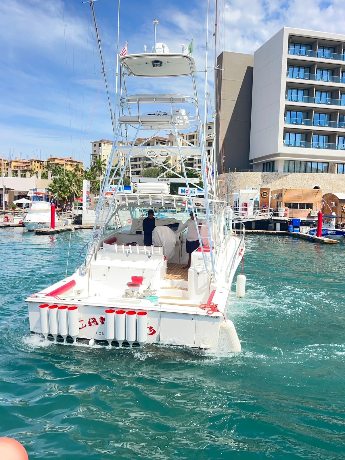 A white fishing boat moving away from the dock, with two people on board, in a marina with modern buildings and other boats.