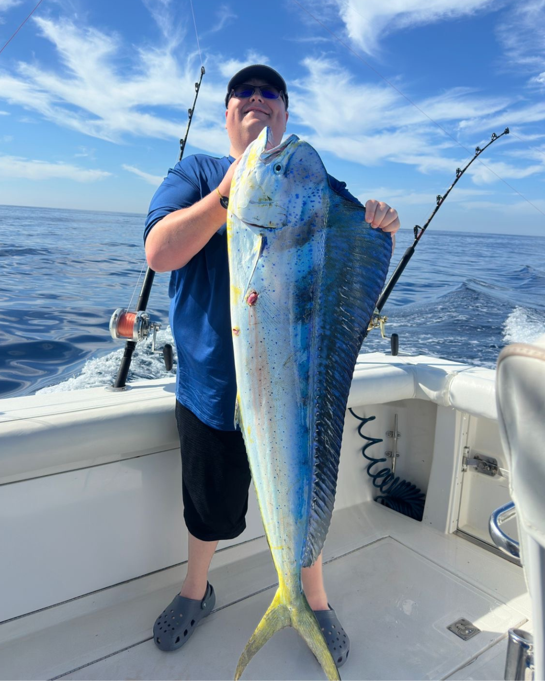 Man holding a large yellowtail fish on a boat with fishing rods, in open water under a blue sky with some clouds.