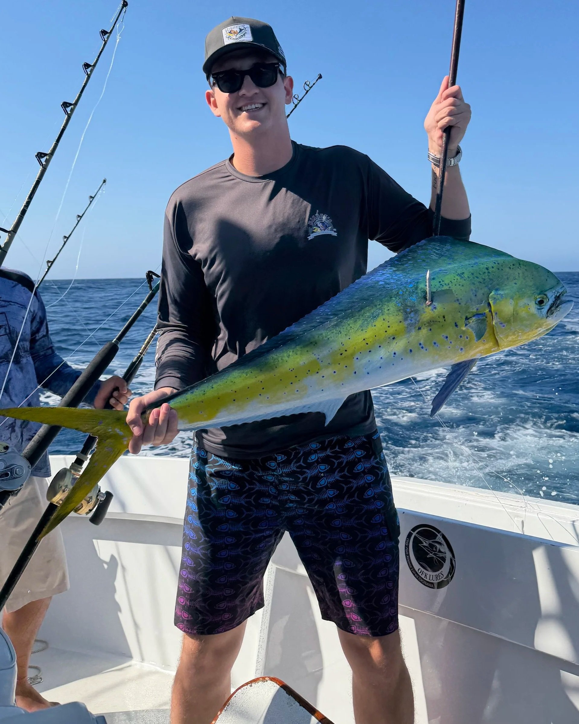 A man wearing sunglasses, a black cap with a logo, and a gray shirt, smiling while holding a large yellow and green fish on a boat. Several fishing rods are visible in the background, with the open ocean and a clear blue sky behind him.