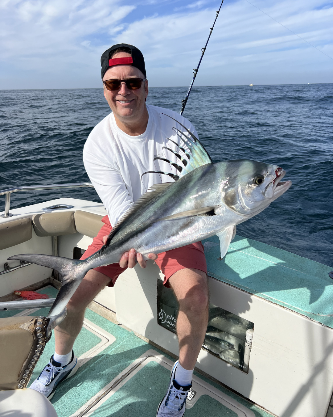 A man on a boat holding a large fish he caught, with the ocean and sky in the background.