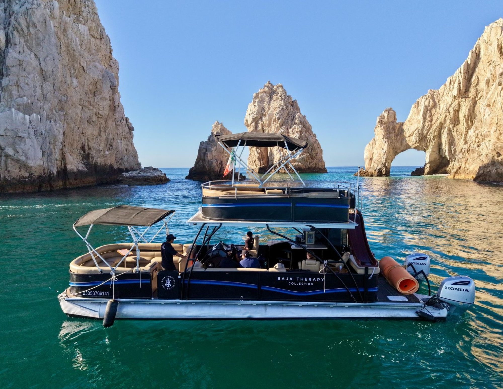 A boat named Baja Therapy Collection with people aboard, floating on turquoise water near rocky cliffs with natural arches and a large rock formation in the background.