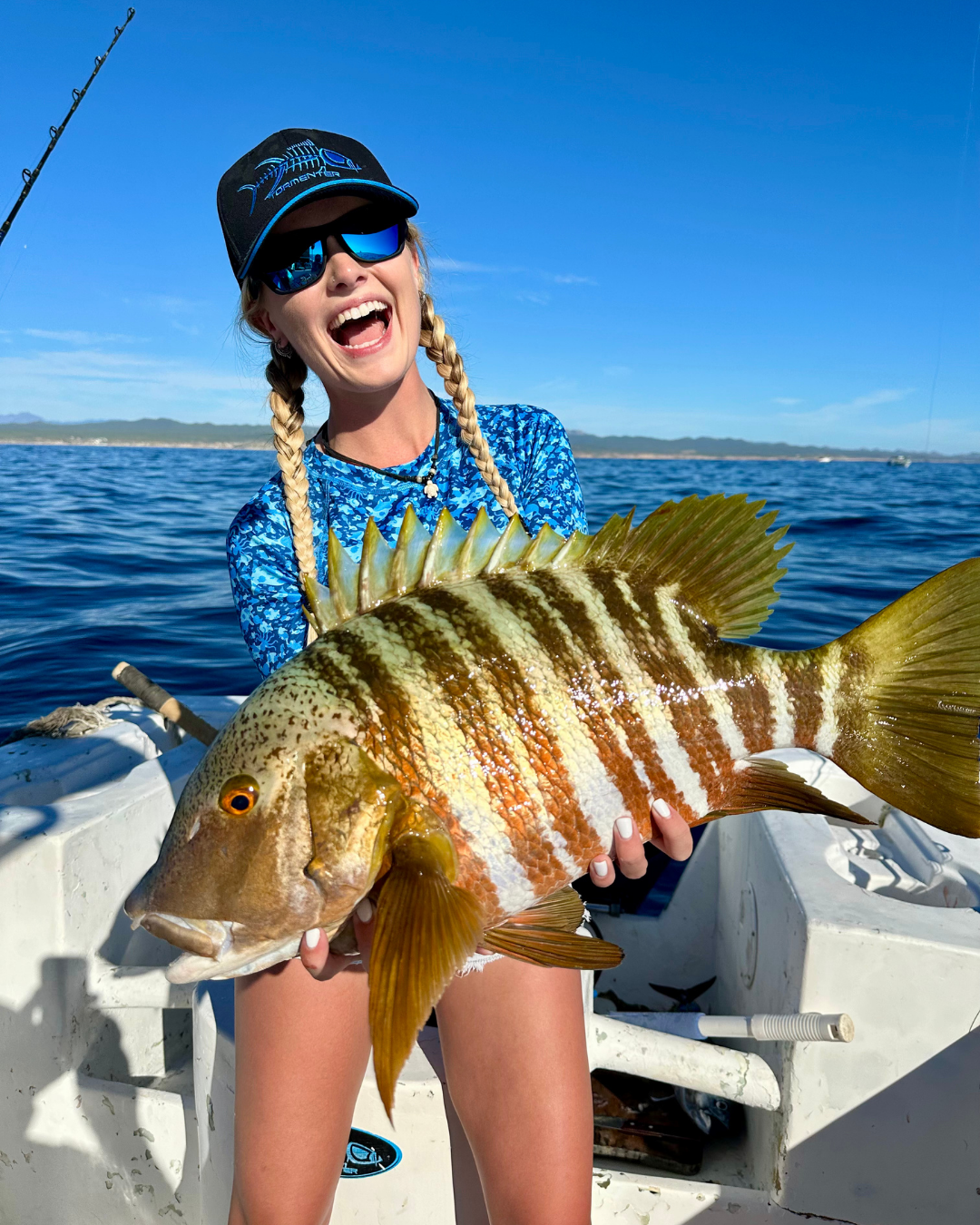Woman holding a large striped fish on a boat in the water, smiling and wearing sunglasses, cap, and a blue patterned shirt.