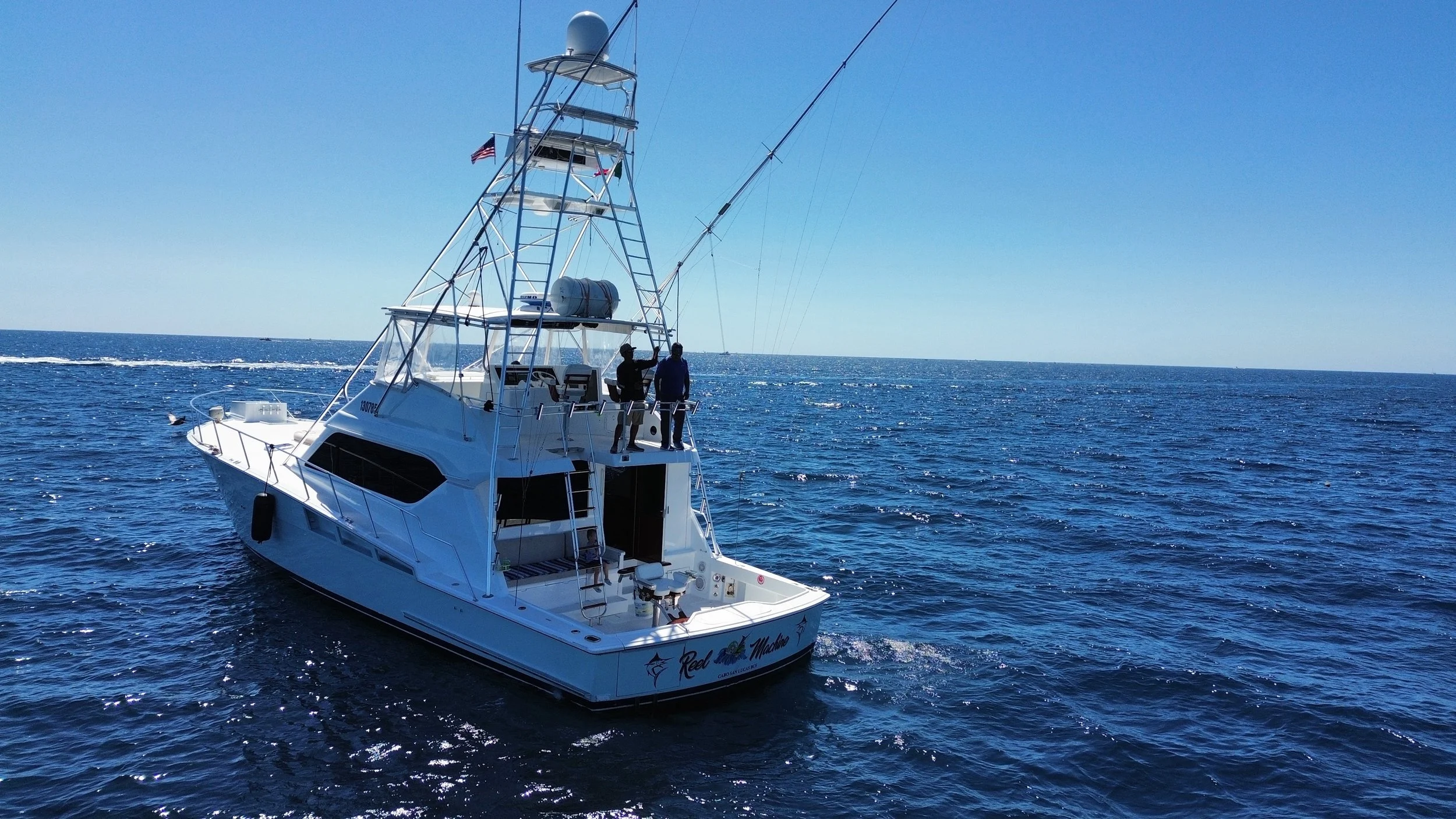 A large white fishing boat named 'Reel Magic' on the open sea under a clear blue sky, with three people on board.
