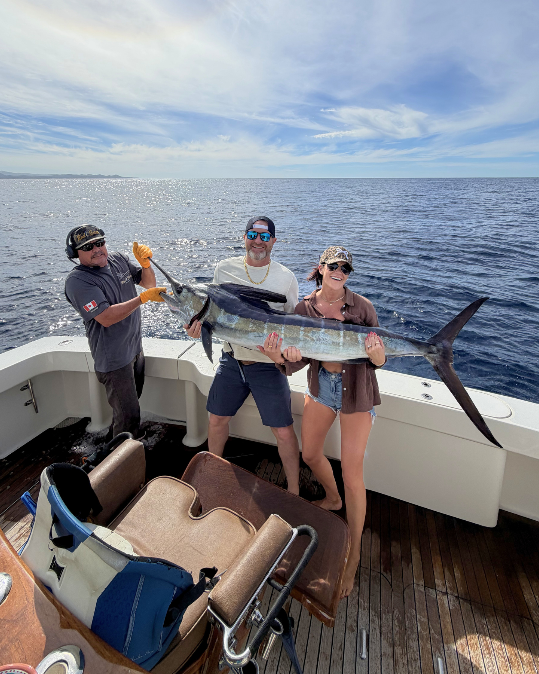 Three people on a boat holding a large fish, a marlin, during daytime with the ocean and sky in the background.