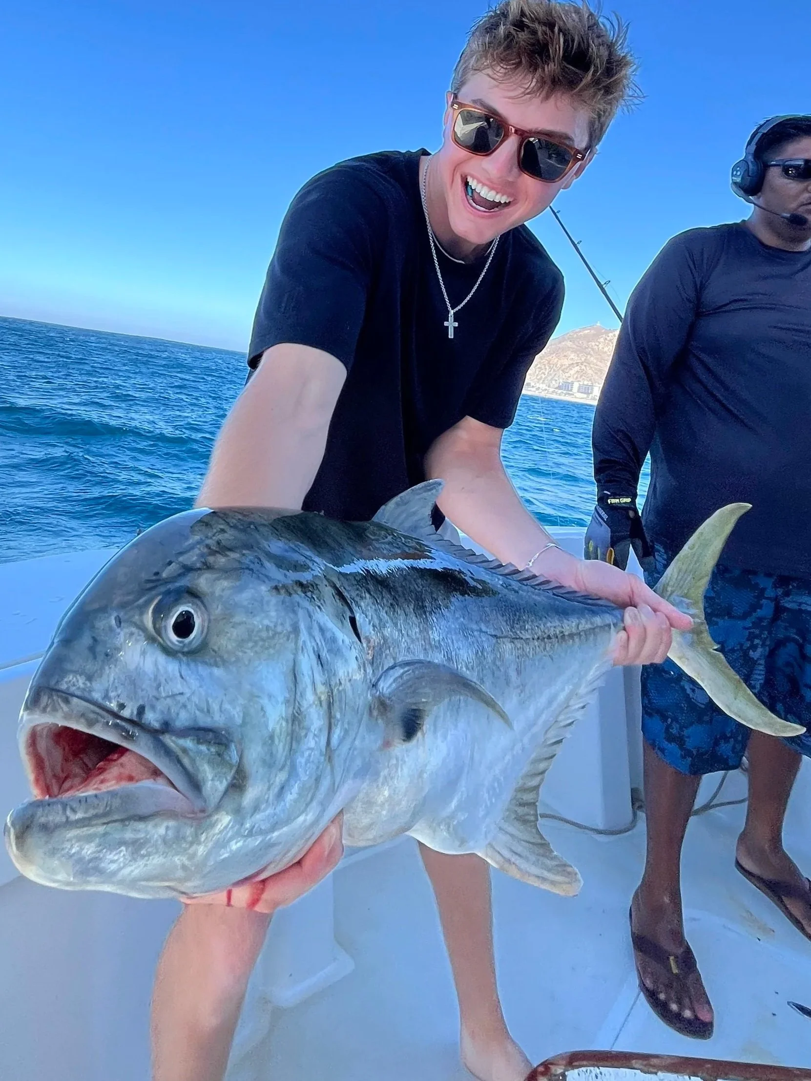 A person holding a large fish on a boat with the ocean and hills in the background, another person in sunglasses with a headset, all under a clear blue sky.