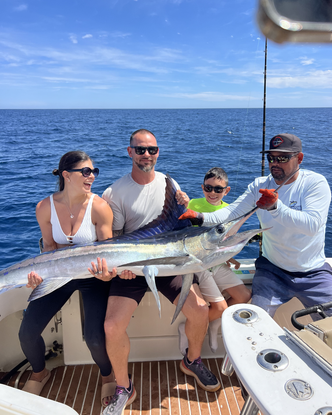 Group of four people on a boat holding a large fish, with the ocean and blue sky in the background.