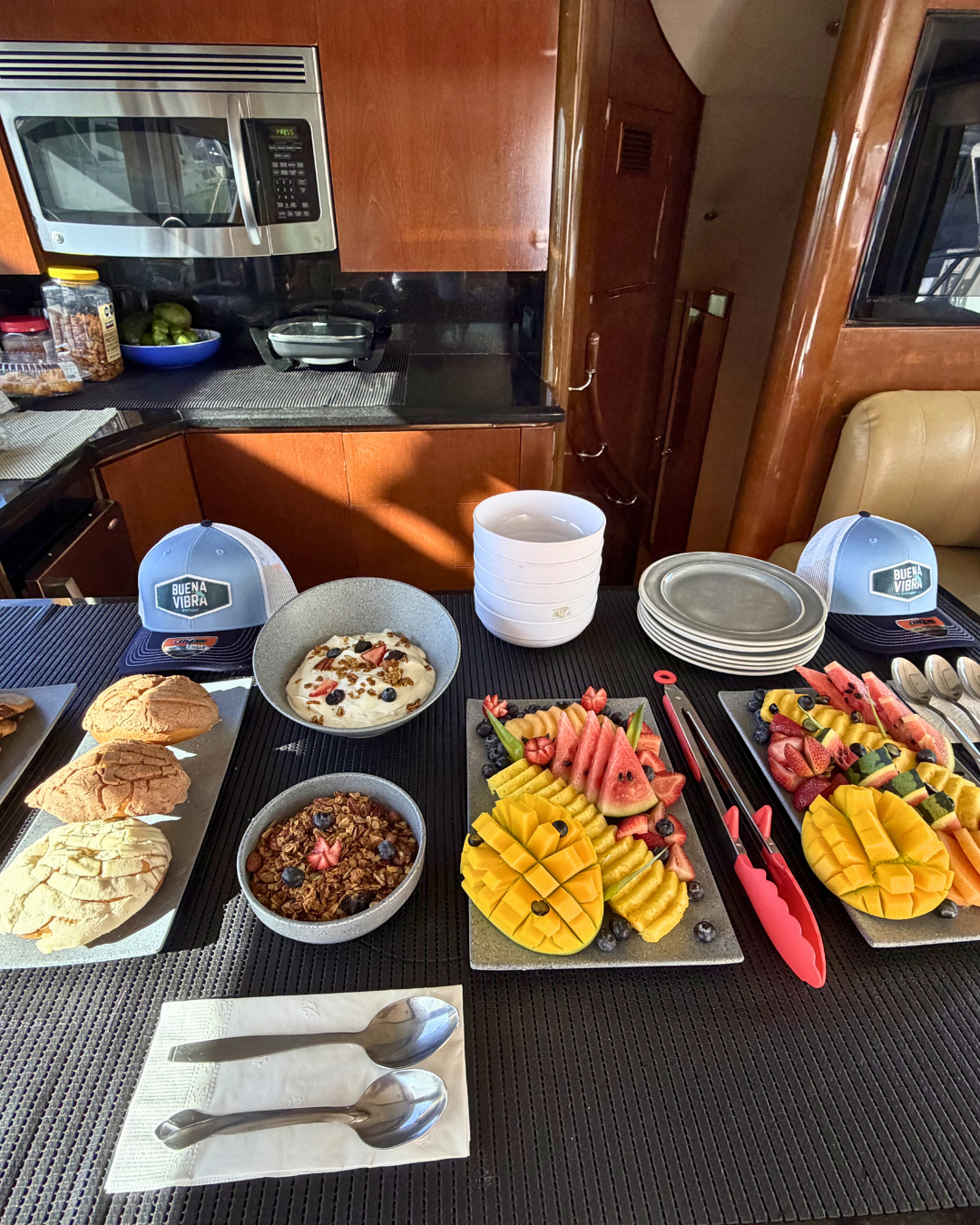 Breakfast spread with bowls of yogurt topped with berries and granola, sliced watermelons and mangos, cookies, plates, and caps on a table in a kitchen setting.