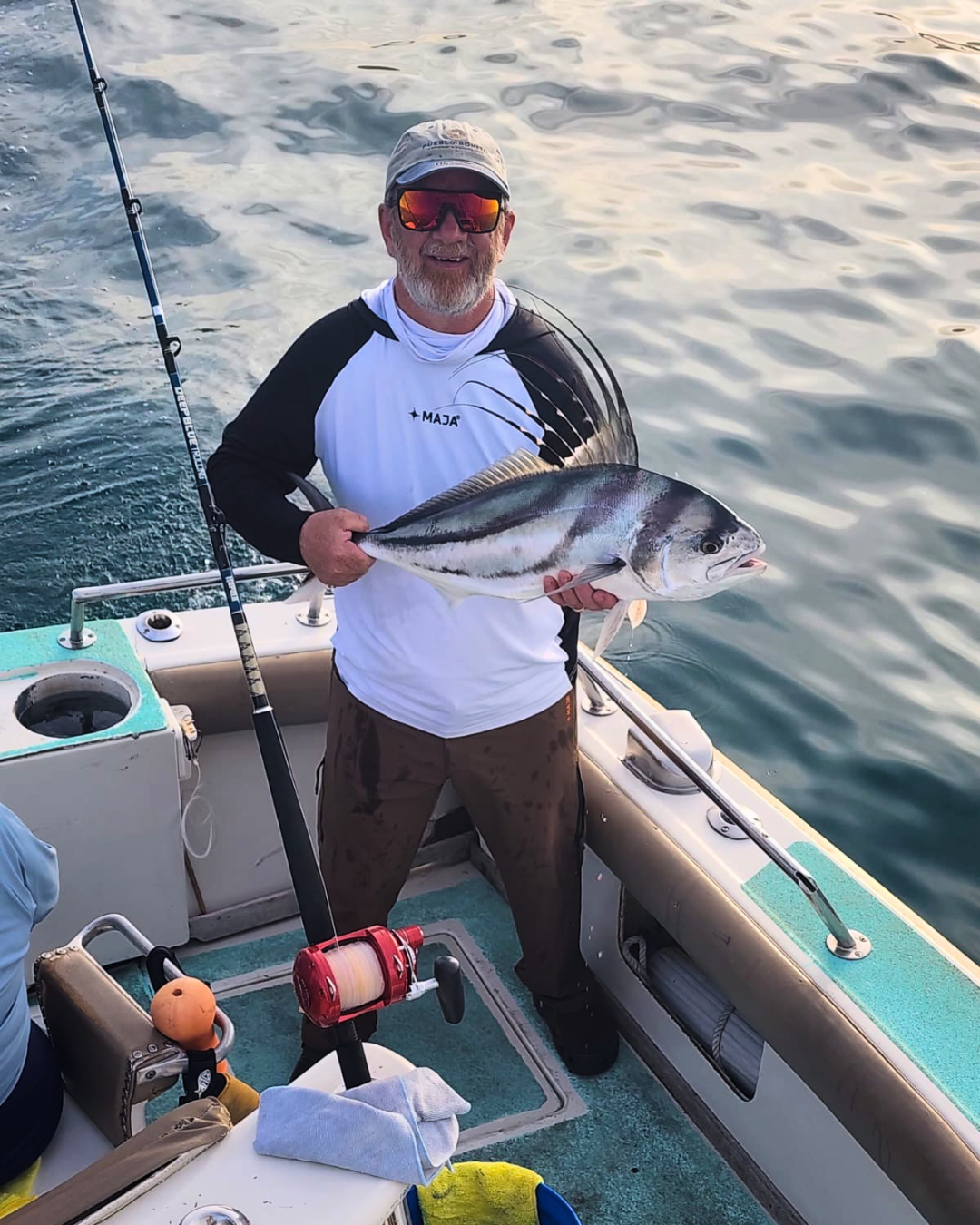 A man on a boat holding a large fish with striped pattern, wearing sunglasses, a baseball cap, a white and black long-sleeve shirt, and brown pants.