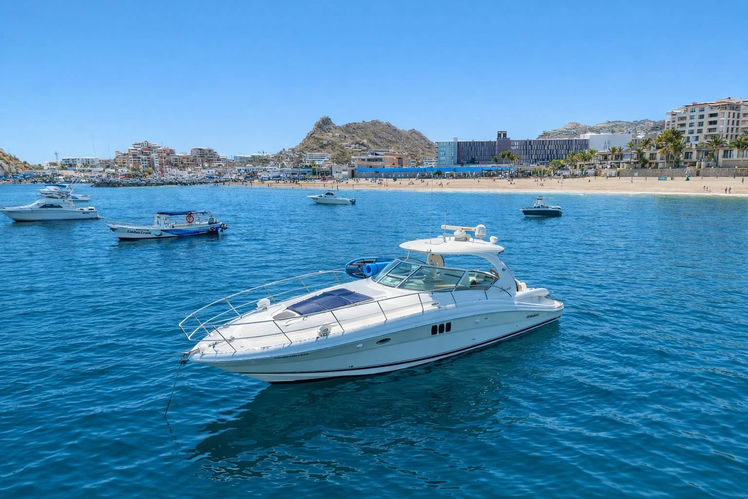 A luxury white yacht docked in a harbor with several other boats on clear blue water, a sandy beach, and buildings with rock formations in the background.