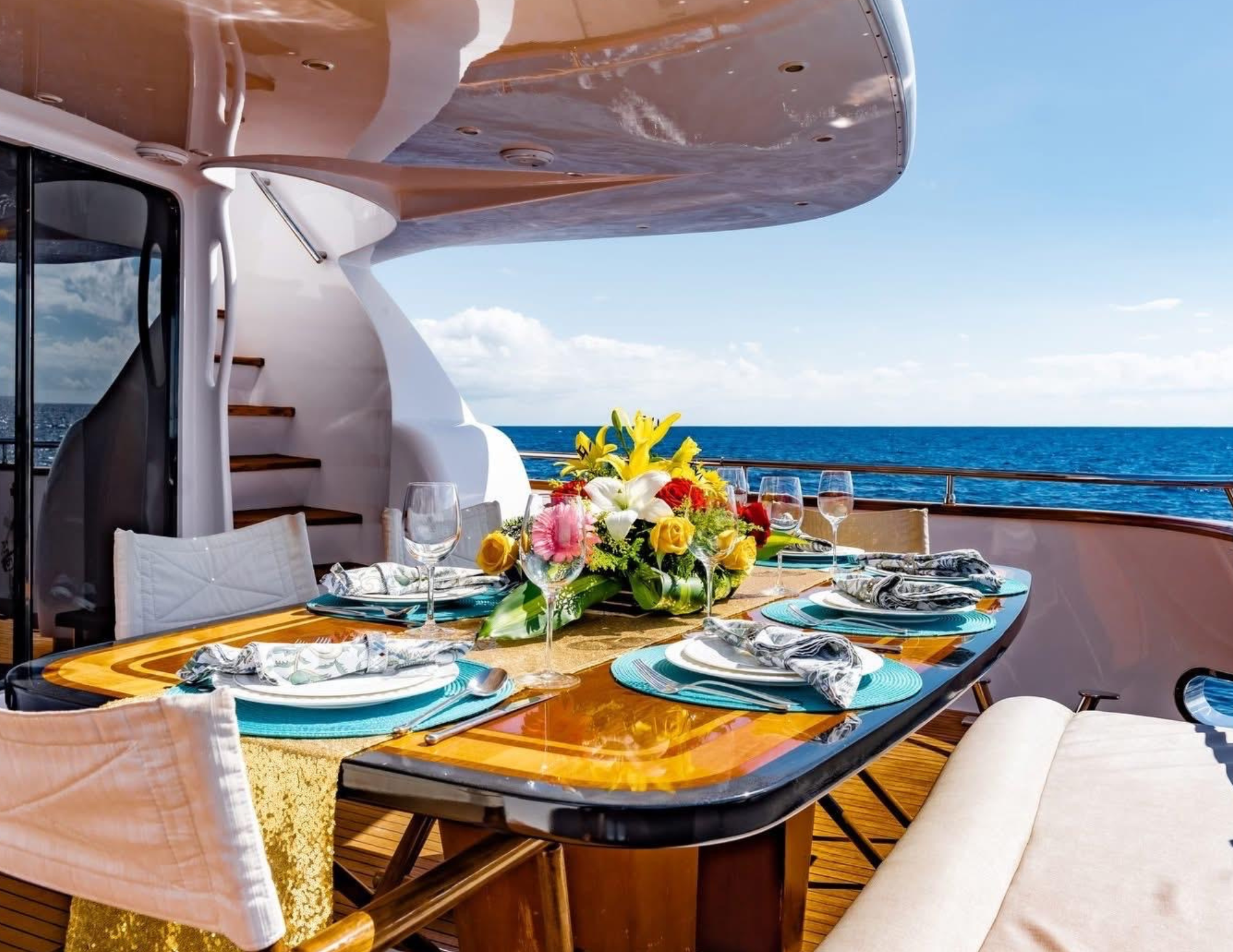 A dining table decorated with a colorful floral centerpiece, set with plates, napkins, and wine glasses on a deck with ocean views, on a yacht.