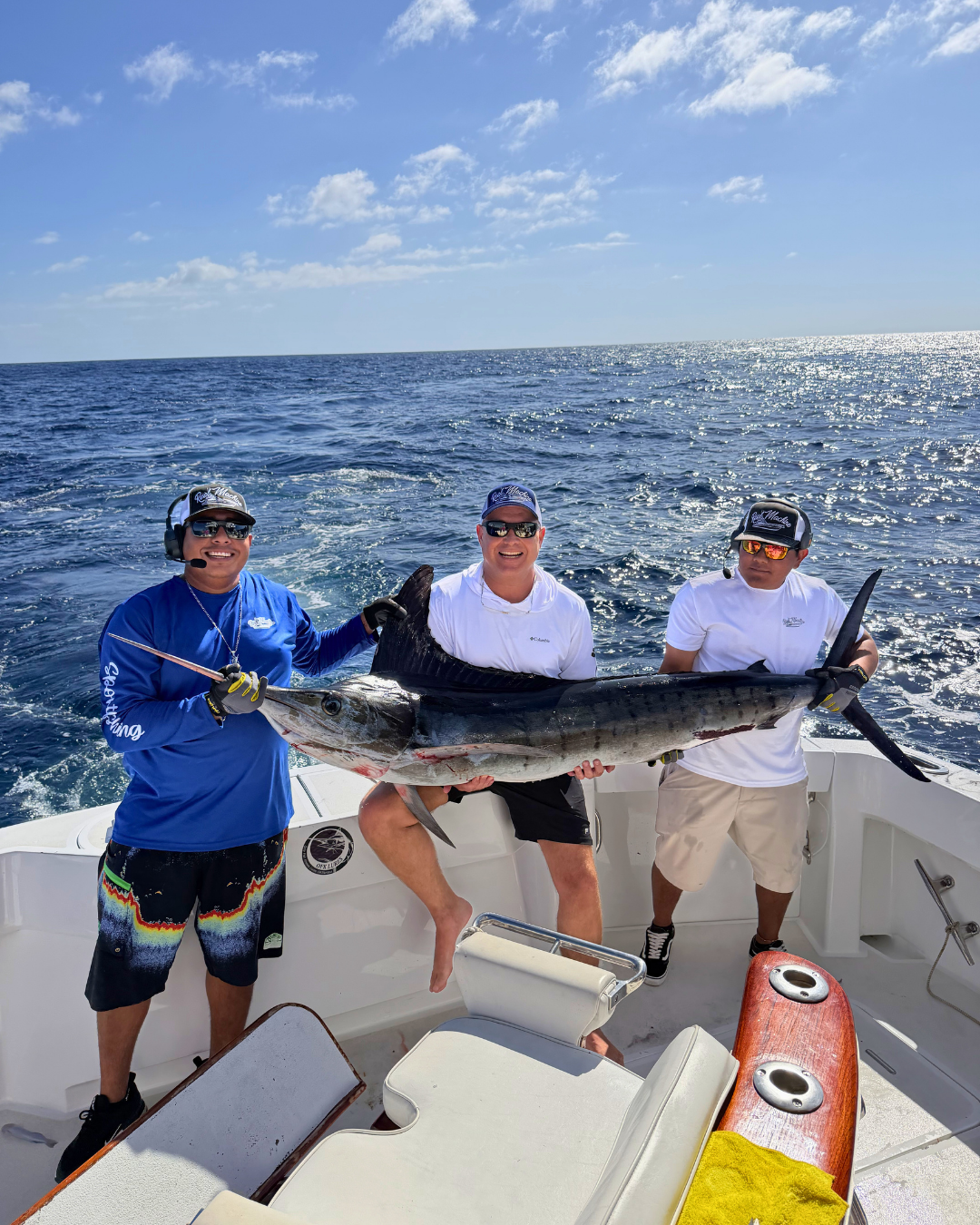 Three men holding a large fish on a boat in the ocean under a blue sky with scattered clouds.