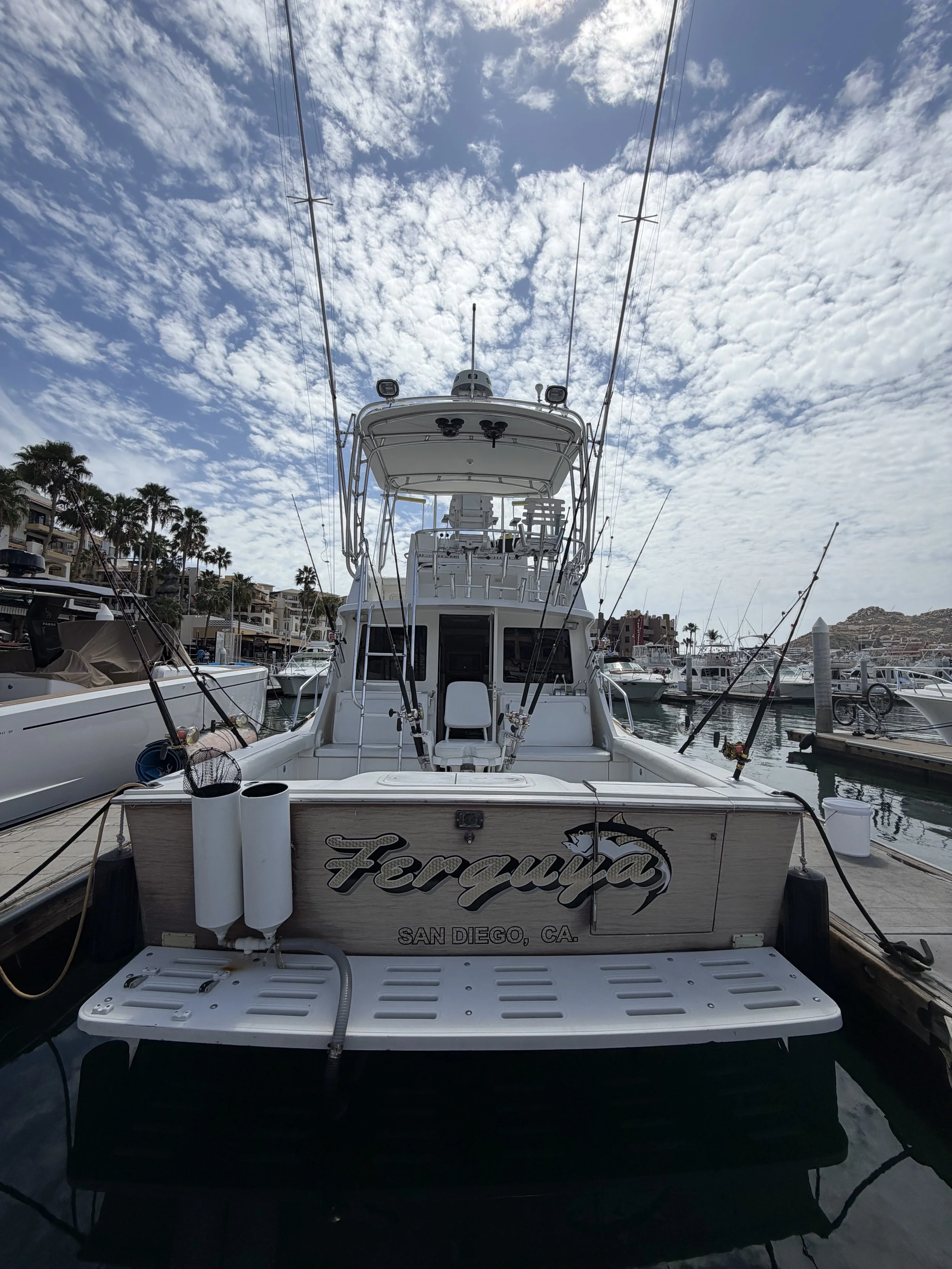 A white fishing boat named Ferquiza docked at a marina in San Diego, California, with multiple fishing rods attached to the boat. The sky is partly cloudy with scattered clouds and blue sky visible.