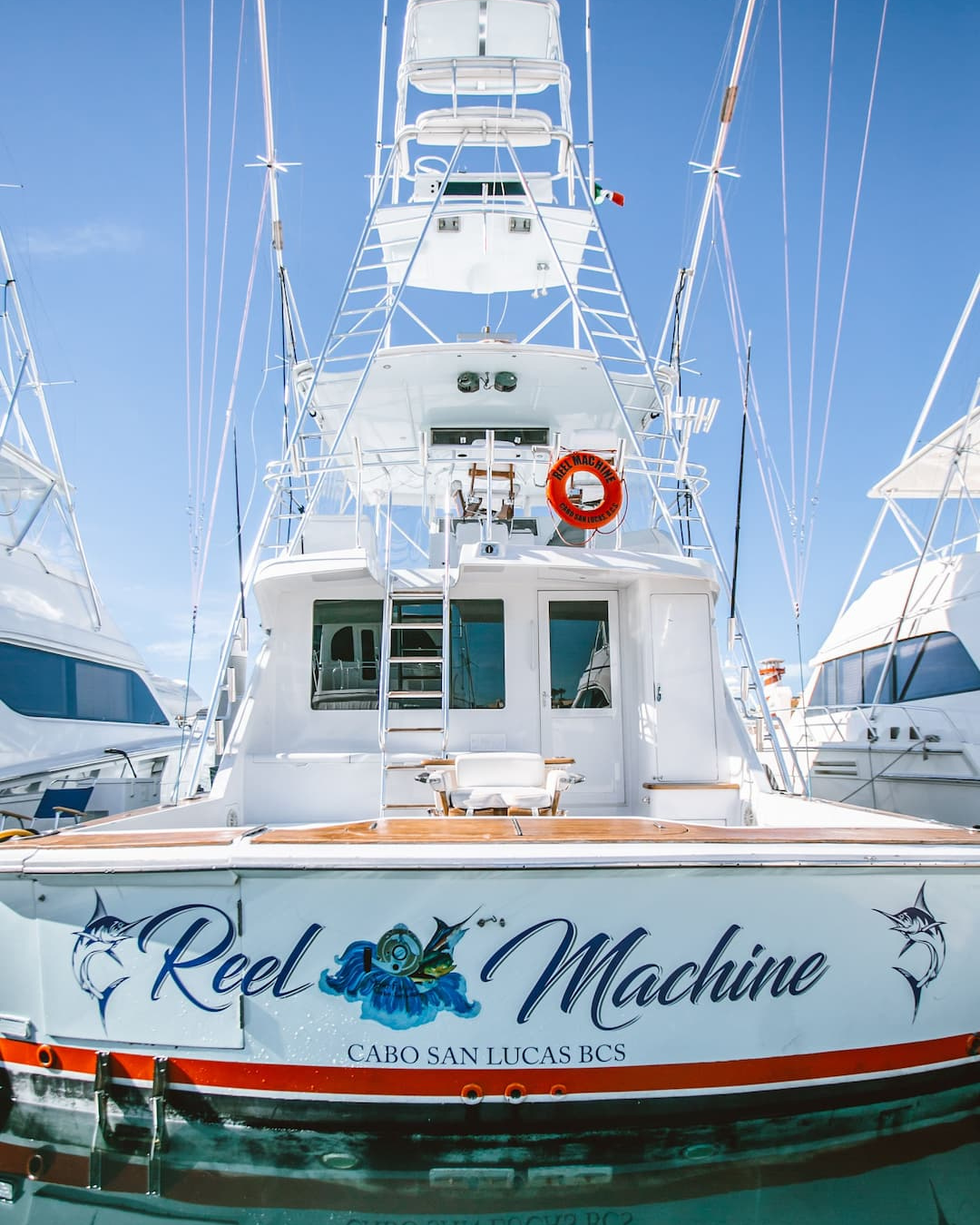 A white fishing yacht named 'Reel Machine' docked at a marina with other boats, under a clear blue sky.