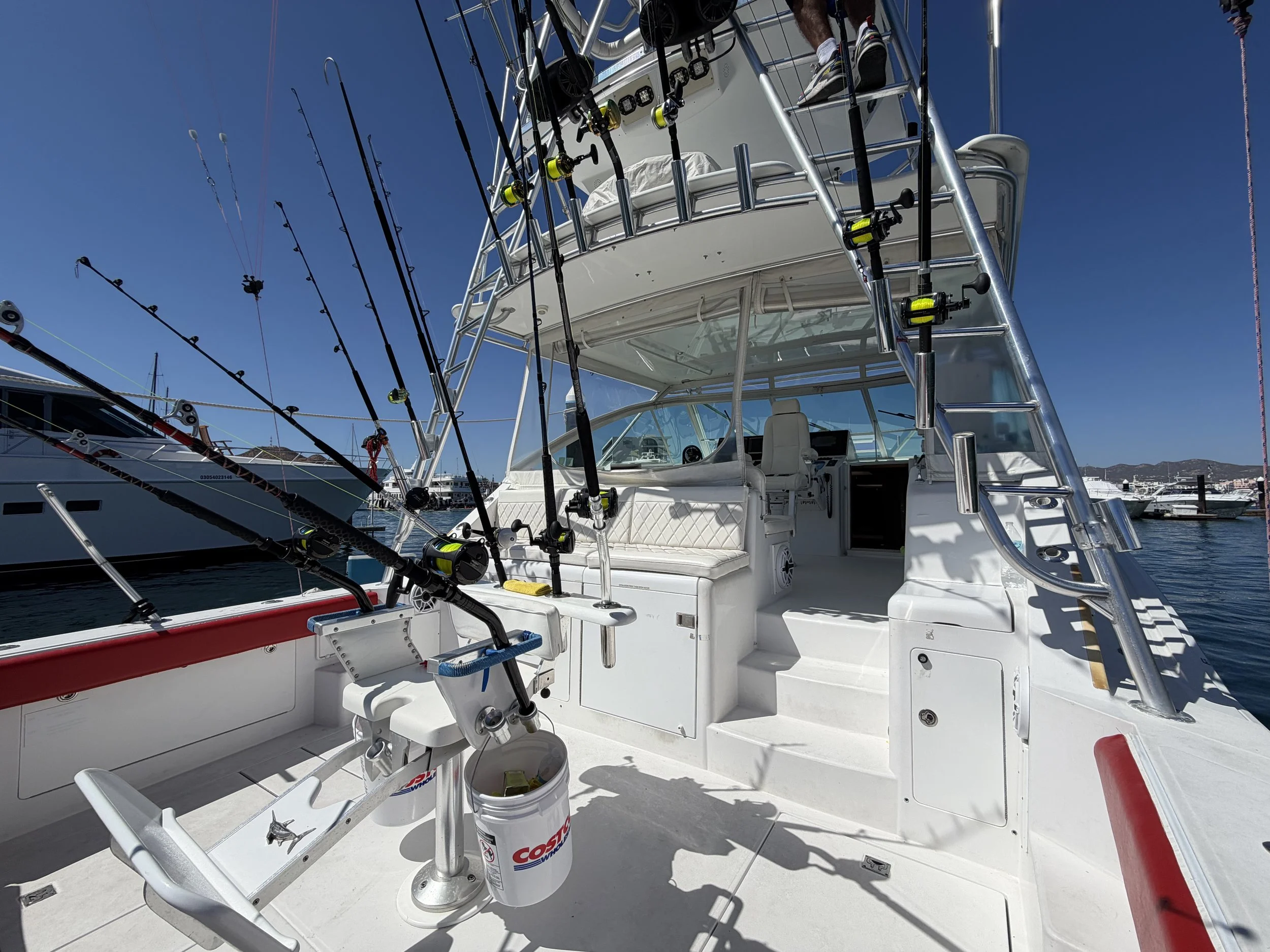A fishing boat with multiple fishing rods mounted on the side, overlooking a marina with other boats under a clear blue sky.