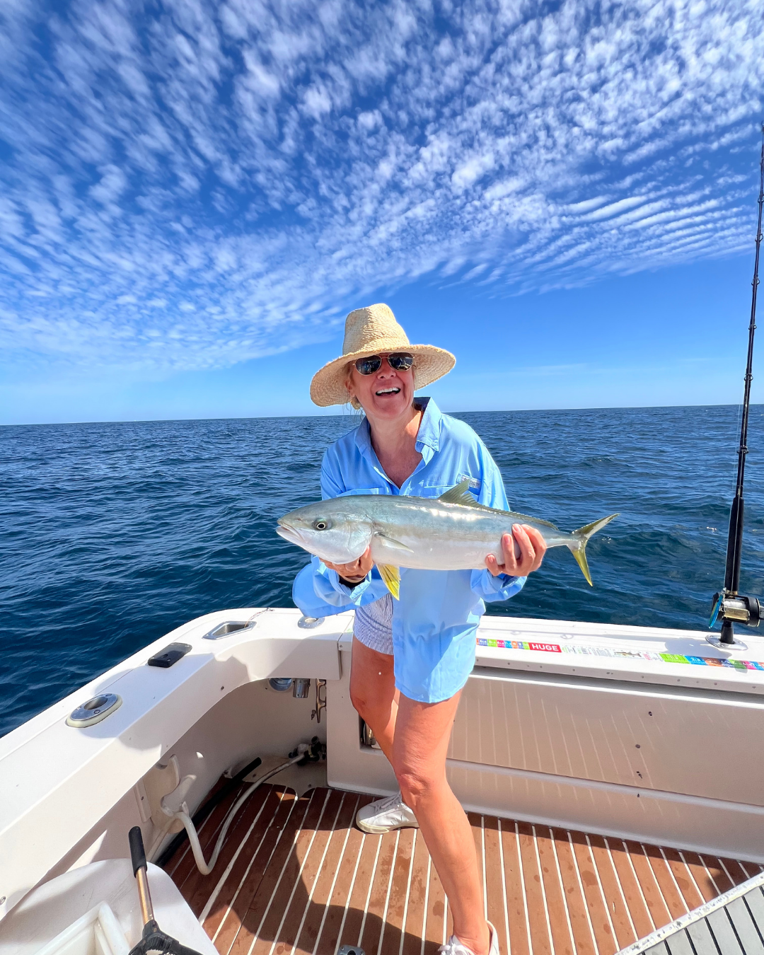 A woman on a boat holding a large fish she caught, wearing a straw hat, sunglasses, light blue shirt, and shorts, with the ocean and a blue sky with scattered clouds in the background.