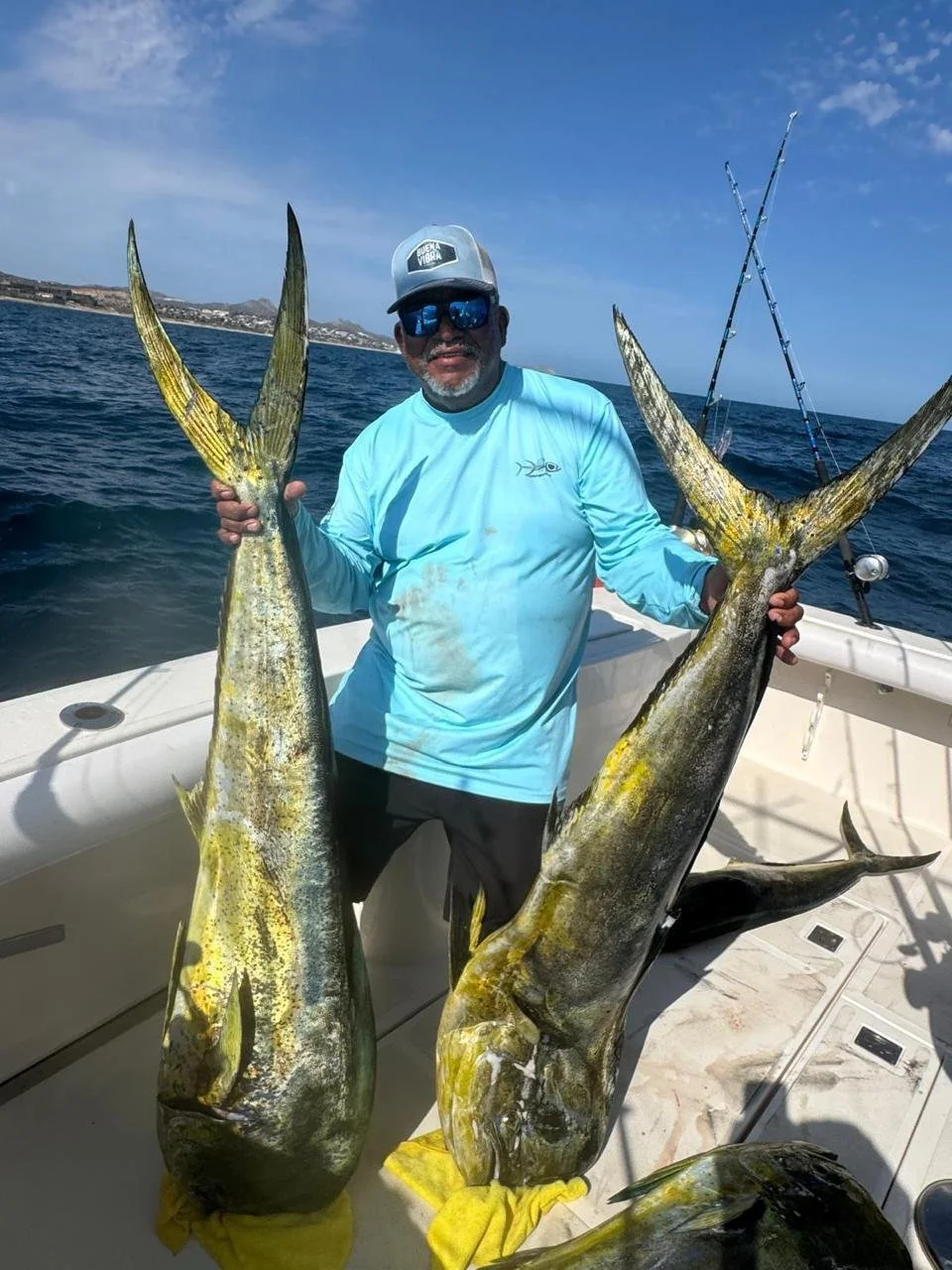 A man on a boat holding two large yellowfin tuna fish, one in each hand, with a body of water and distant shoreline in the background.