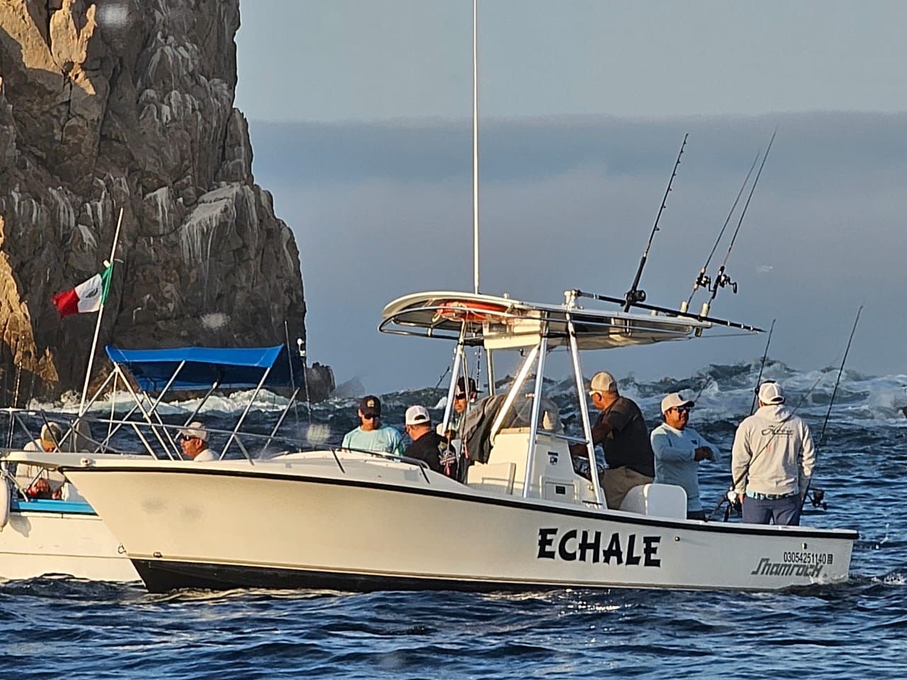A group of people fishing on a boat named 'Echale' nearby a rocky coastline with a Mexican flag, with fishermen holding fishing rods in the water.