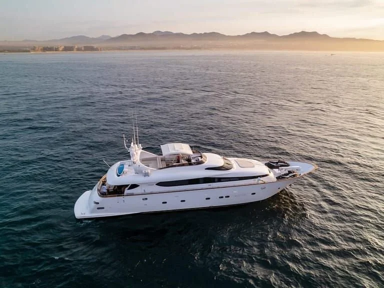 A luxury white yacht sailing on calm ocean waters near a coastline with mountains in the background during sunset.