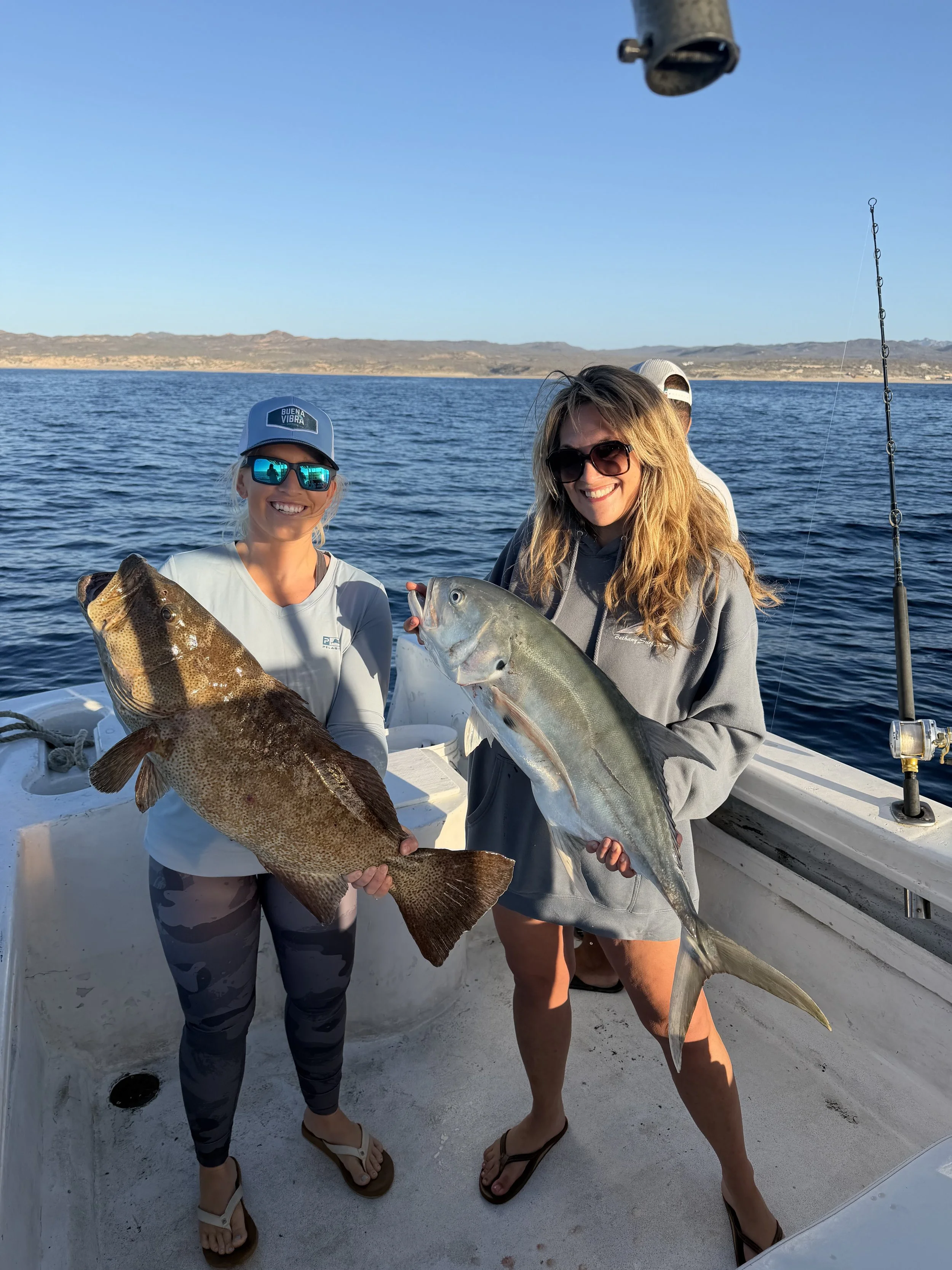 Two women on a boat holding large fish they caught, with a body of water and distant mountains in the background.