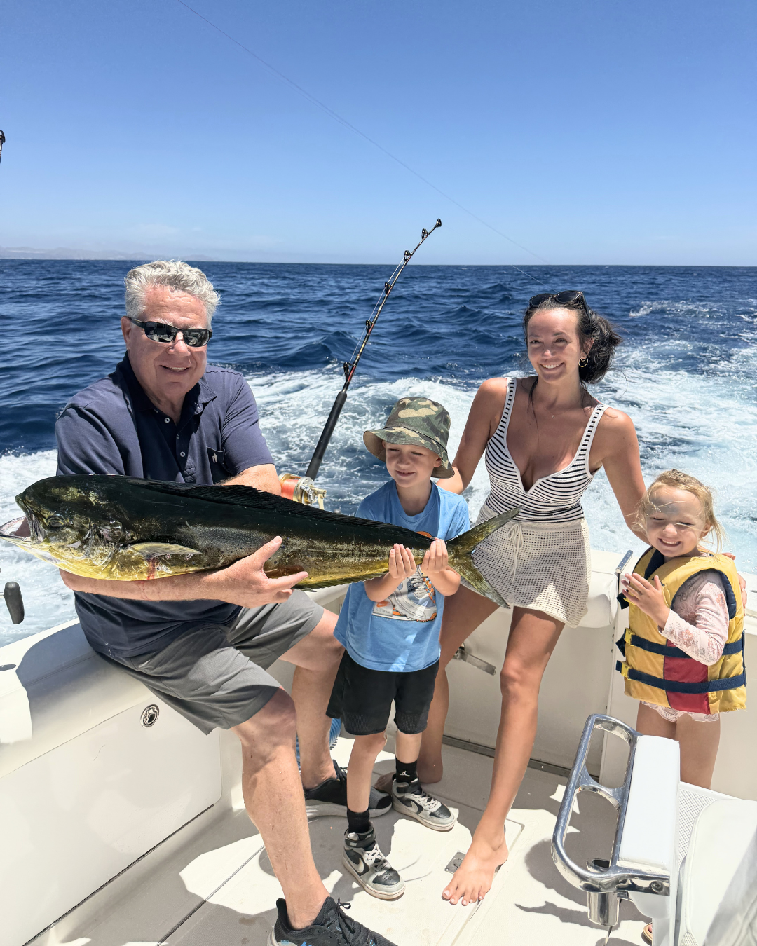 A man and a woman on a boat holding a large fish, with two children smiling nearby, in the open ocean under a clear blue sky.