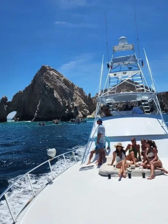 Group of people relaxing on a yacht near rocky coastline with large rock arch formation in the background, sunny day with clear blue sky.