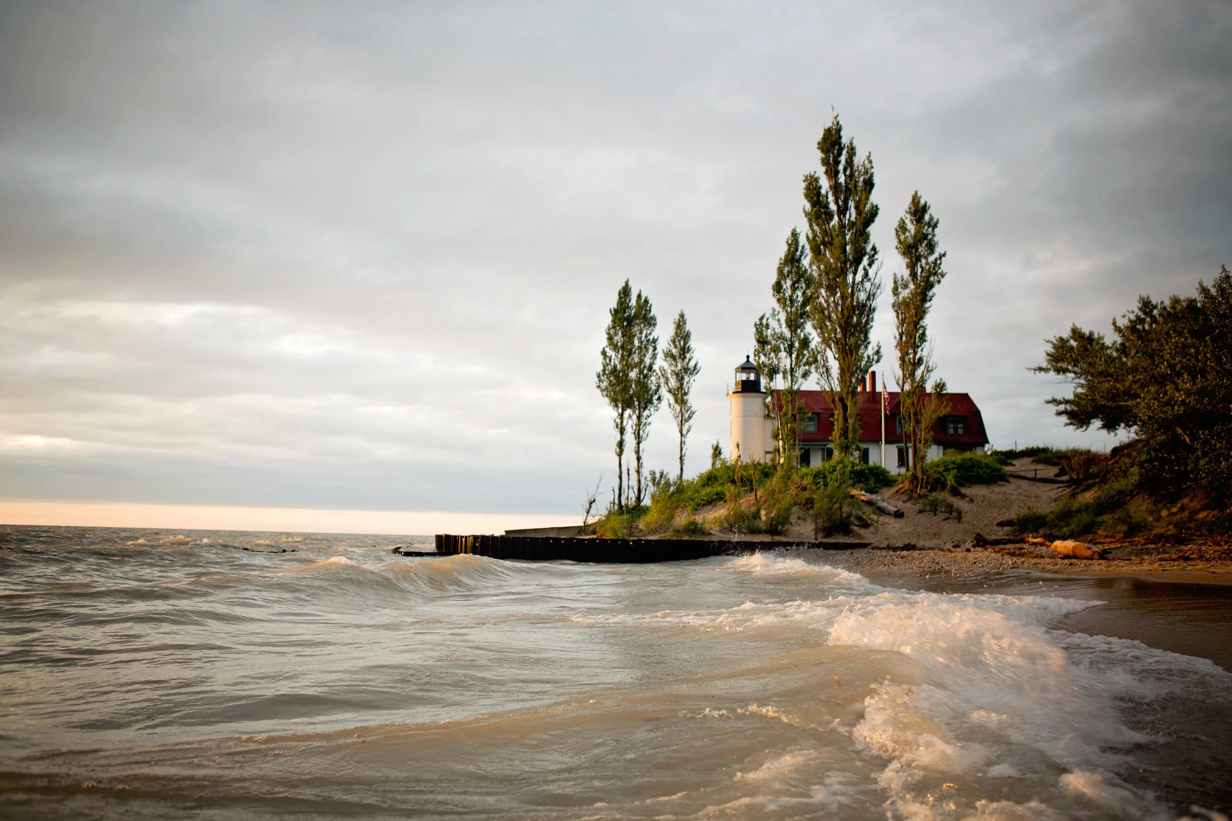 A lighthouse and house on a hill overlooking the ocean, with trees and a cloudy sky in the background.