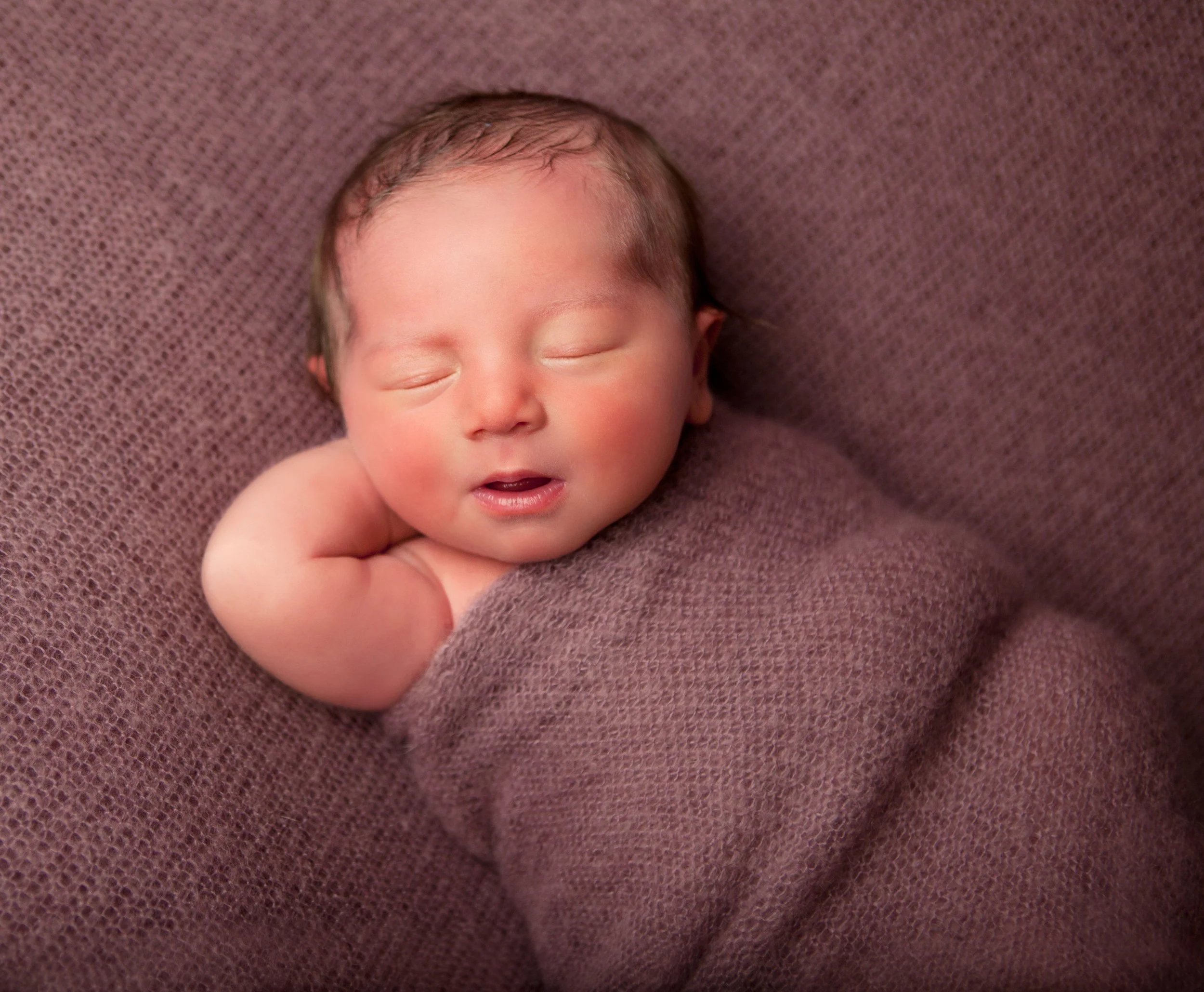 Close-up of a sleeping newborn baby with fair skin, tiny features, and light brown hair, lying on a textured mauve fabric surface.