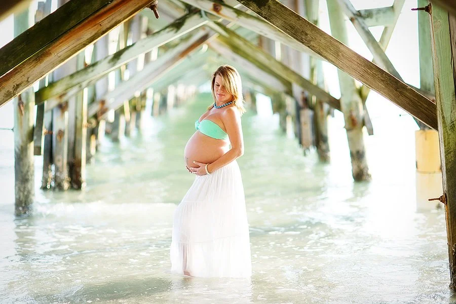 A pregnant woman in a green bikini top and white skirt standing in shallow water under a wooden pier with sunlight filtering through.