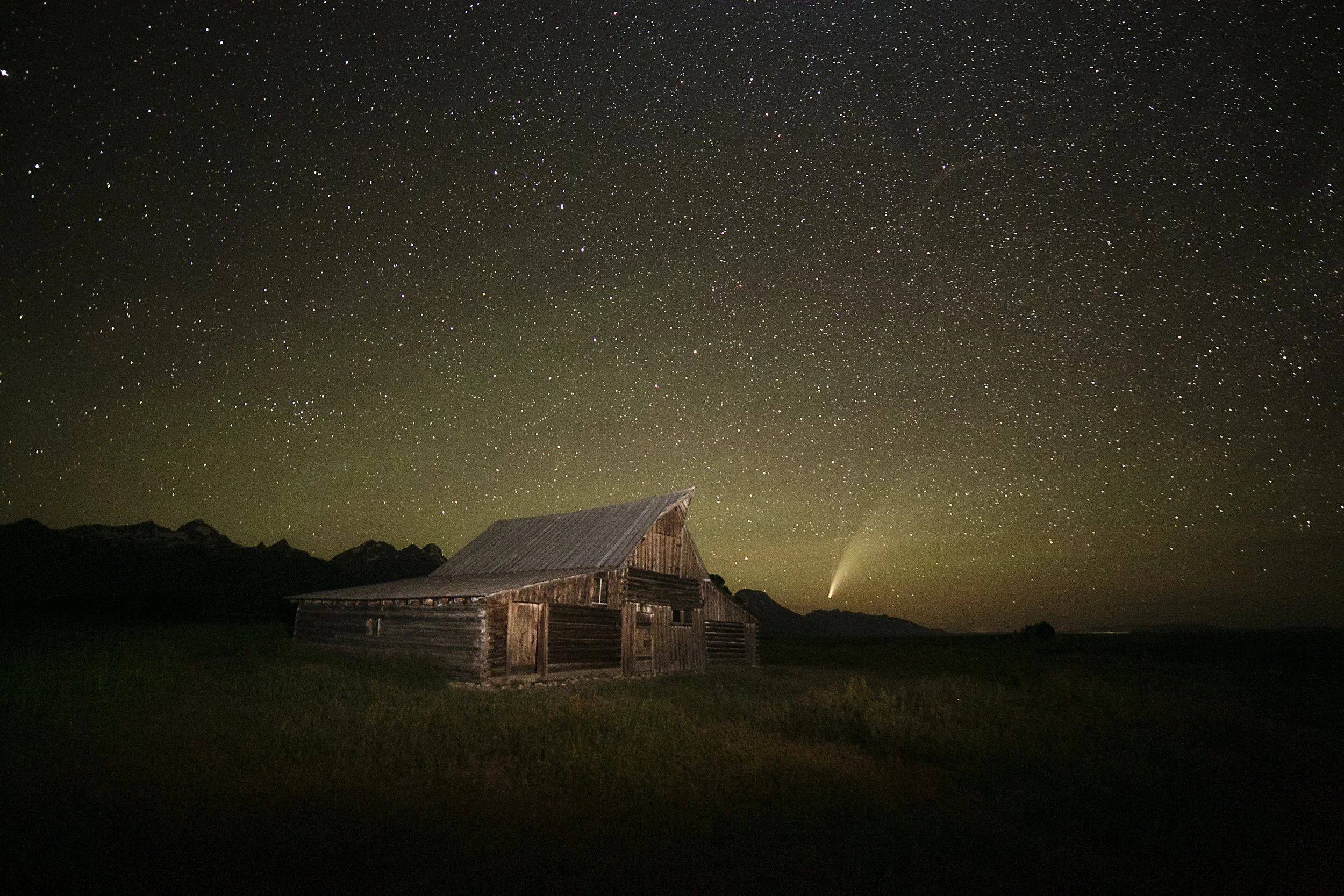A starry night sky over a rustic wooden barn with mountains in the background and a bright comet or shooting star visible near the horizon.
